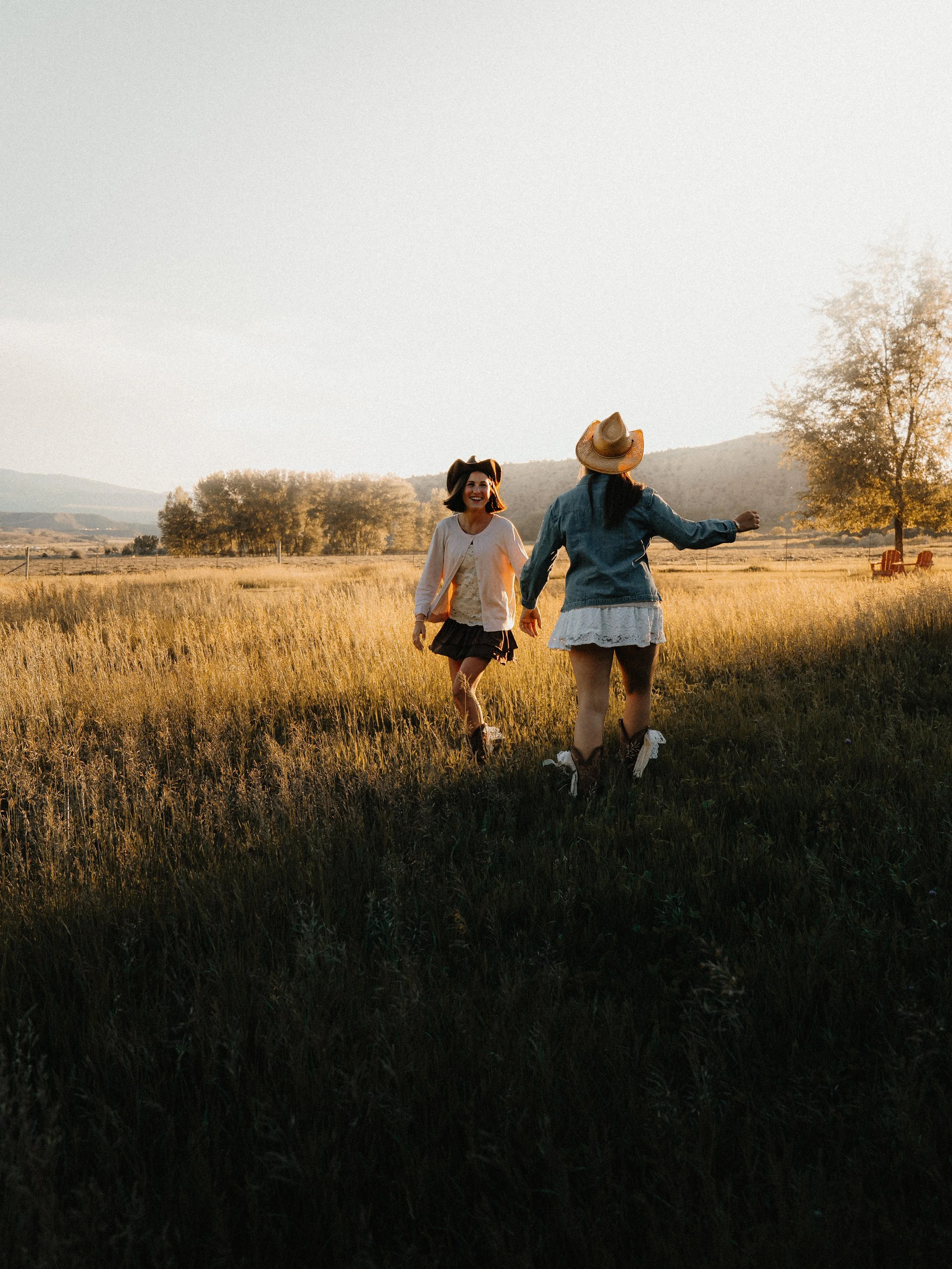 Two women walking through a grassy field during sunset, smiling and enjoying each other's company.