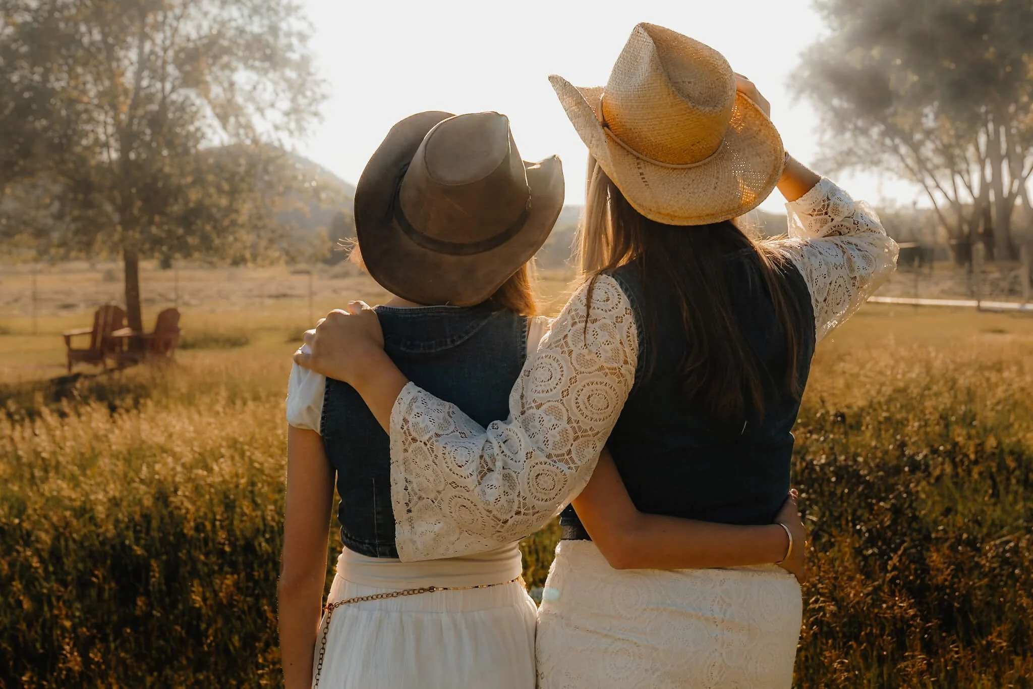 Two women wearing hats and lace tops, standing close together in a field during sunset, with trees and chairs in the background.