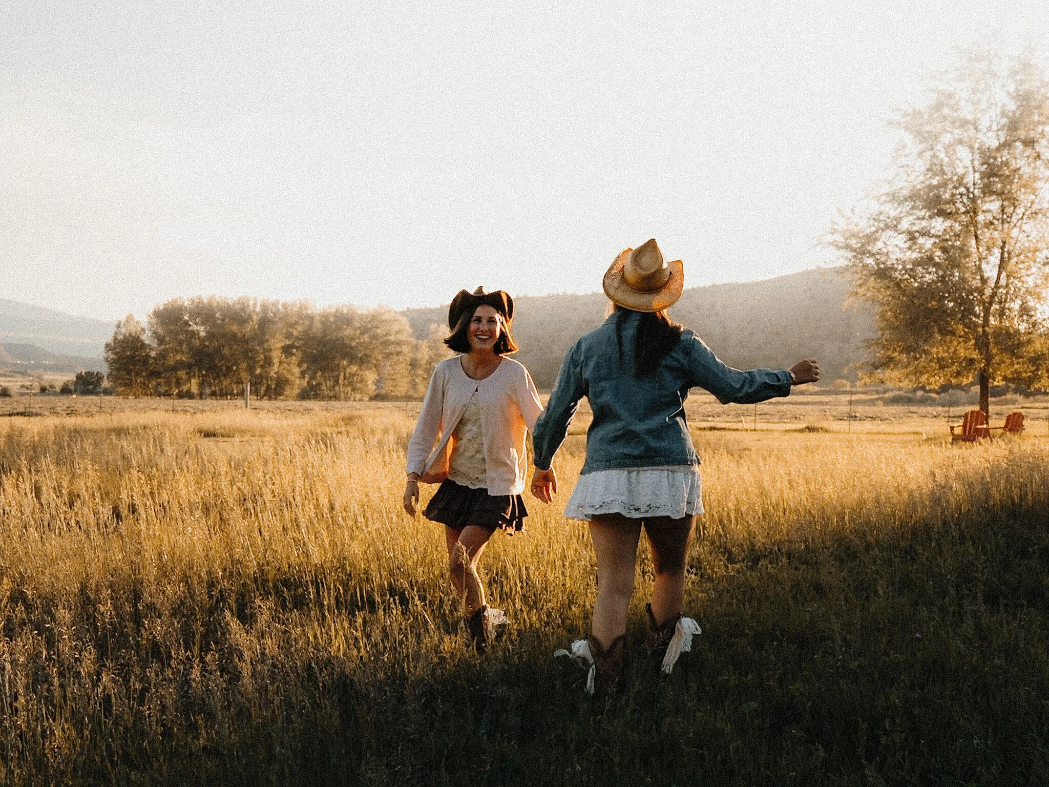 Two women in cowboy hats holding hands and running in a grassy field during sunset, with trees and distant mountains in the background.