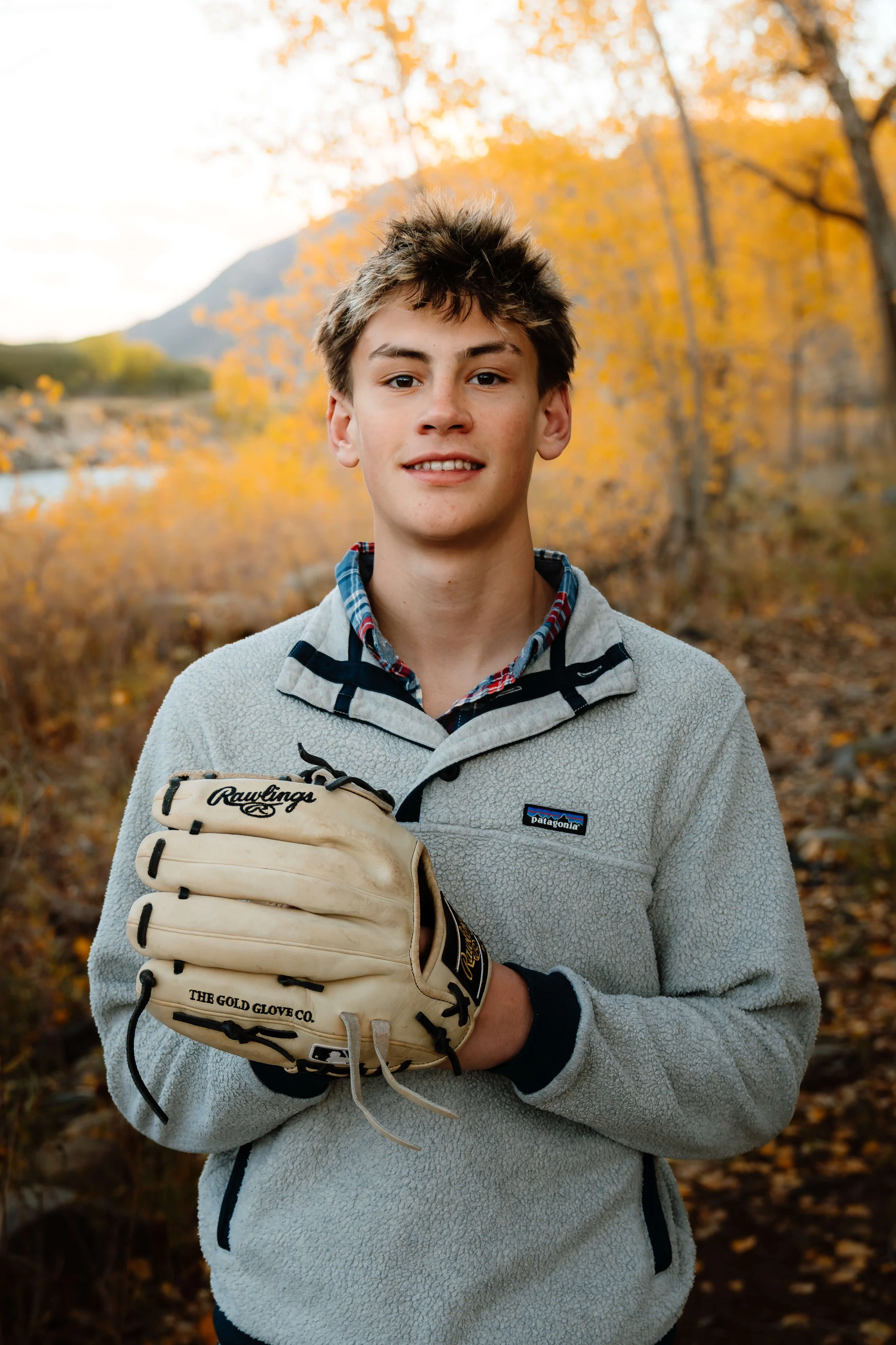 A teenage boy standing outdoors in a fall landscape, holding a baseball glove, wearing a gray Patagonia fleece jacket and a plaid shirt underneath.