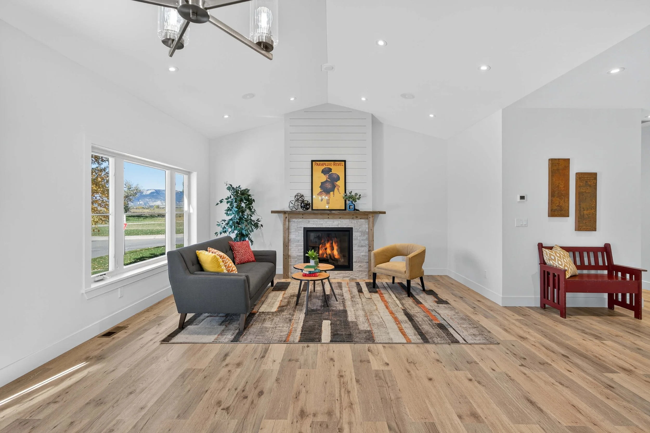 Living room with white walls, wooden floor, window on the left, fireplace in the center with a painting above, gray sofa with yellow and red pillows, yellow armchair, red bench, and colorful rug.