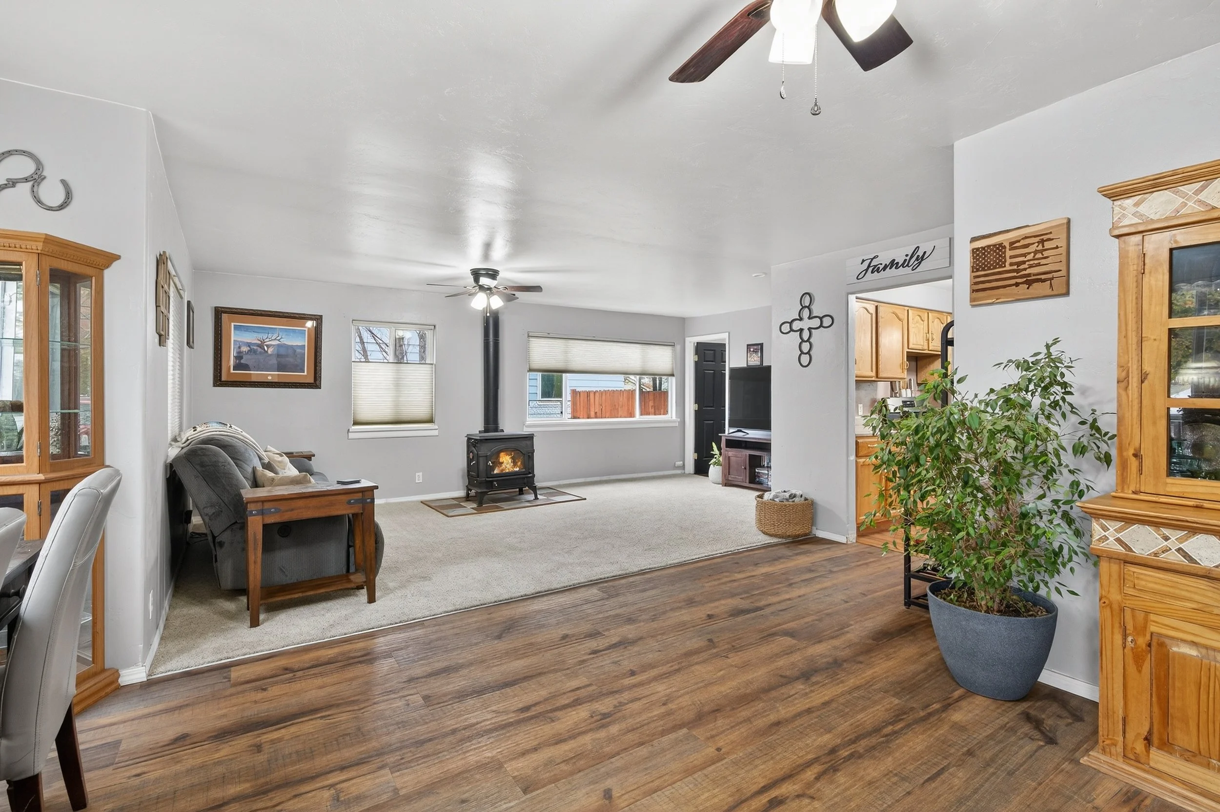 Living room with wooden flooring, white walls, a fireplace, large windows with blinds, and a ceiling fan with lights.