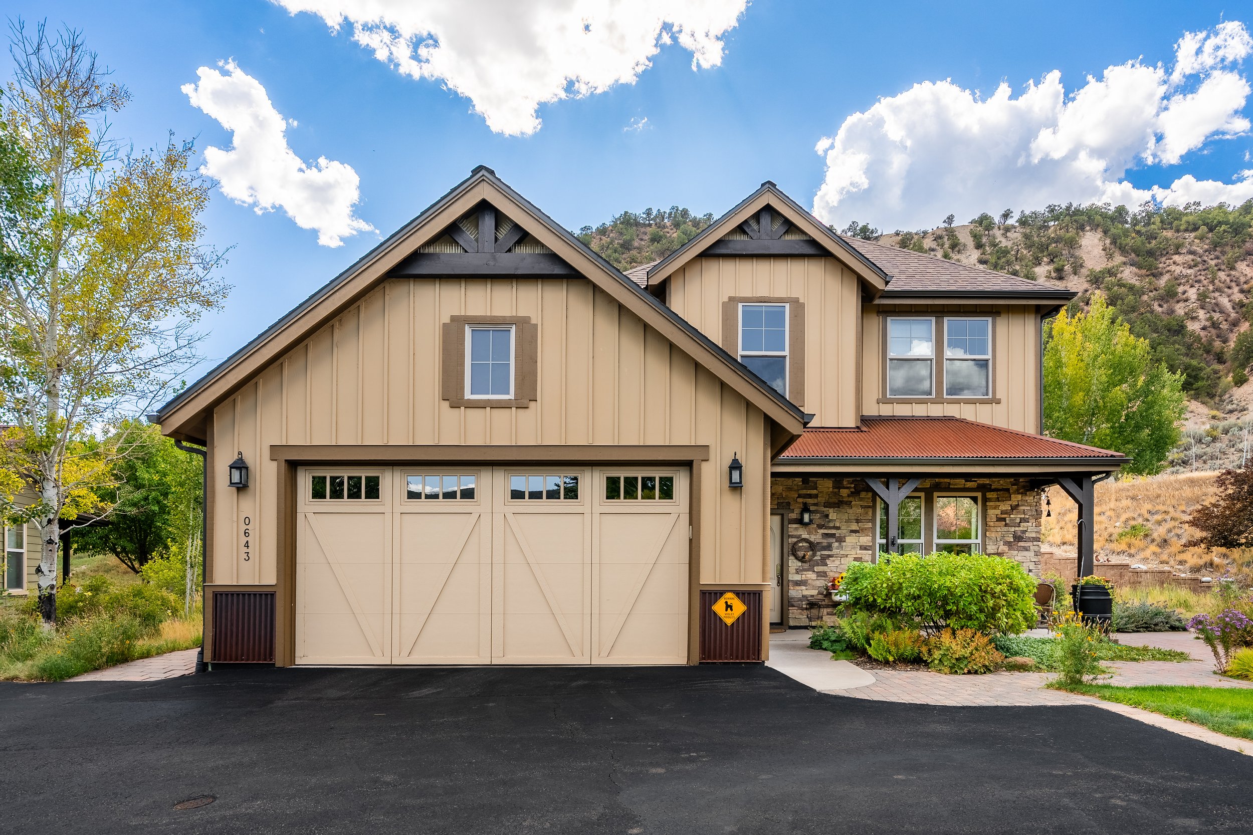 A beige two-story house with a garage, trees, and mountain scenery under a blue sky with clouds.