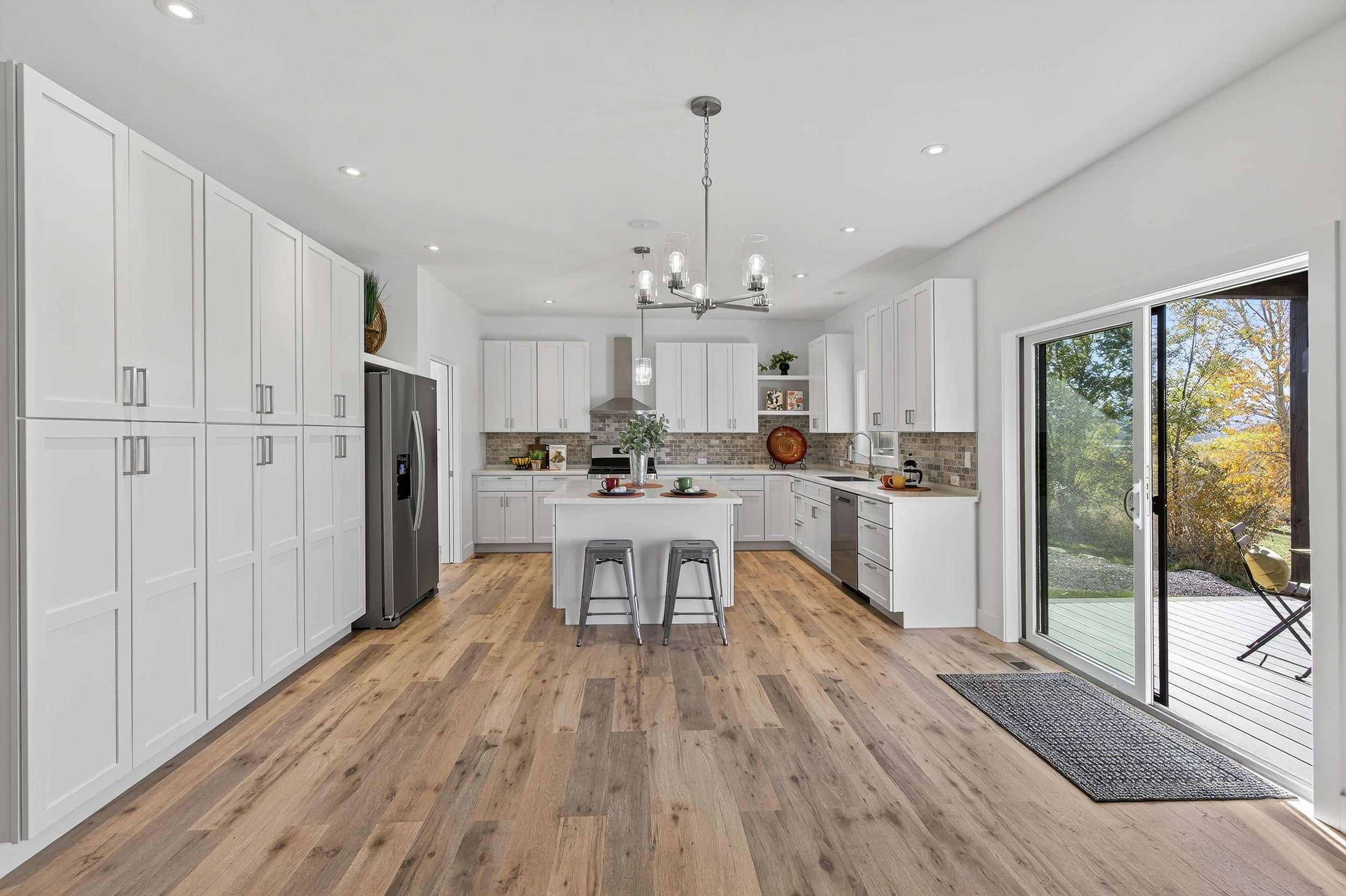 Bright modern kitchen with white cabinets, stainless steel refrigerator, center island with decor, hardwood floors, sliding glass door leading to outdoor patio with outdoor chair, and large window showing trees outside.