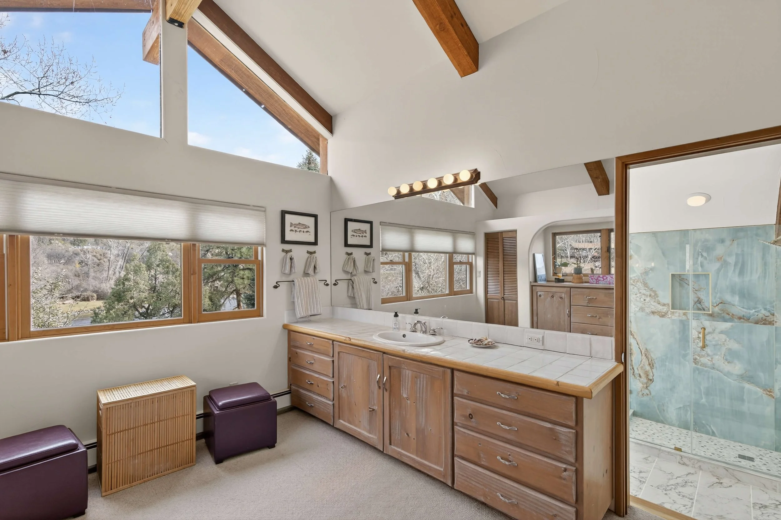 A bathroom with a wooden vanity, a white tiled countertop, a sink, and a large mirror above. There are windows with blinds, wooden beams on the ceiling, and a glass shower with blue marble tiles.
