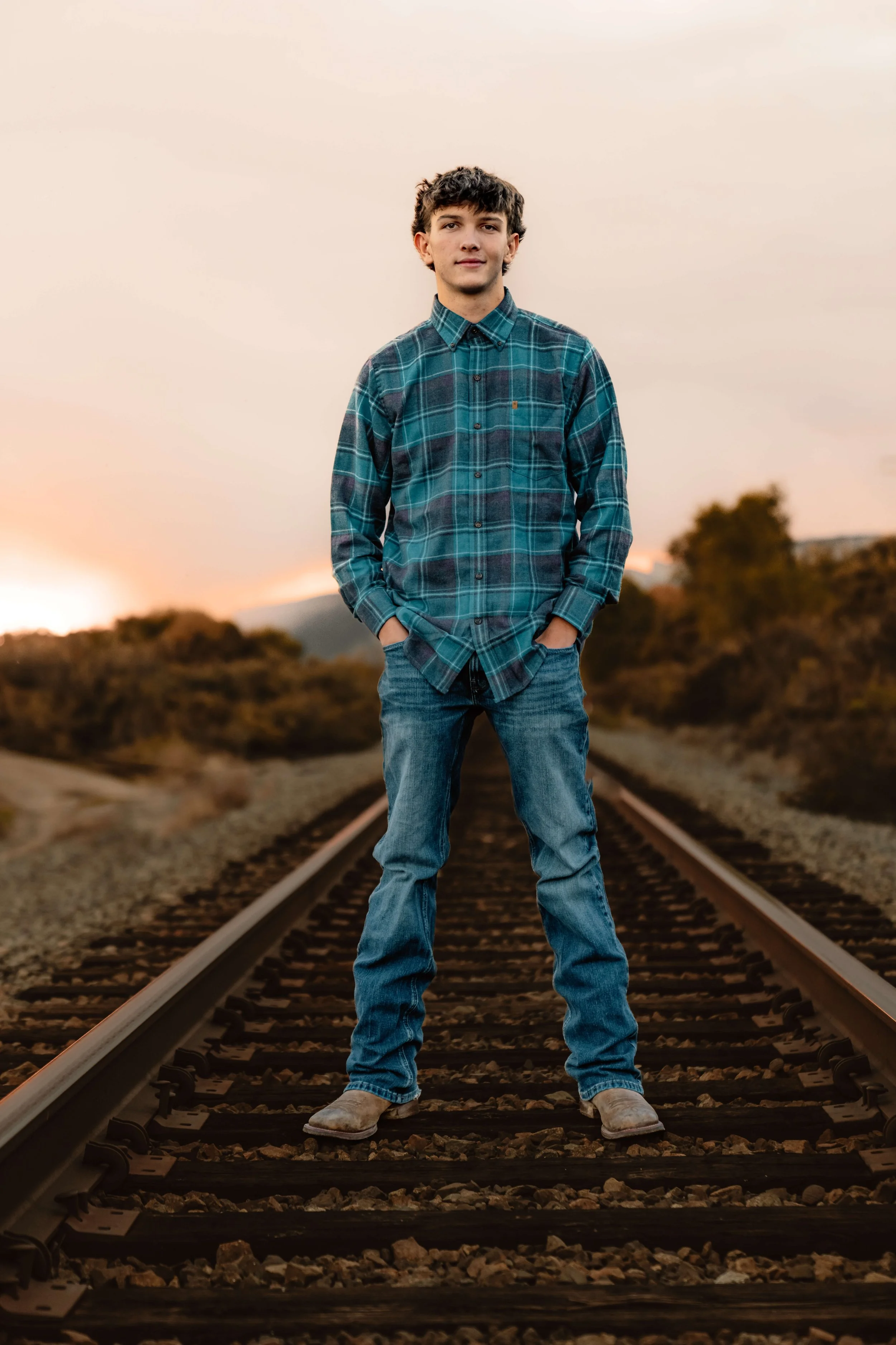 A young man standing on railway tracks during sunset, wearing a blue plaid shirt, jeans, and boots.