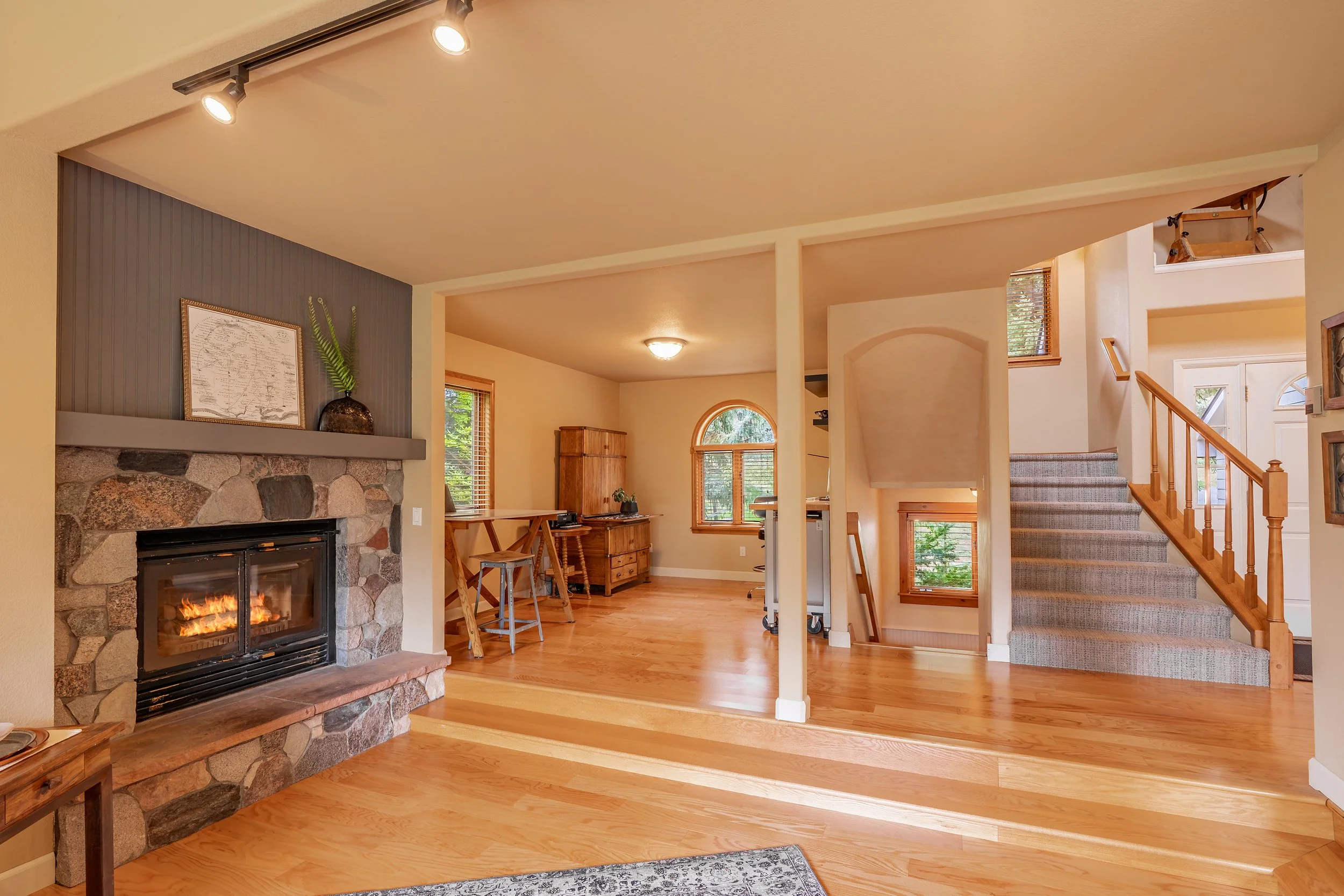 Living room with stone fireplace, hardwood floors, staircase with carpet, and windows with wooden trims.