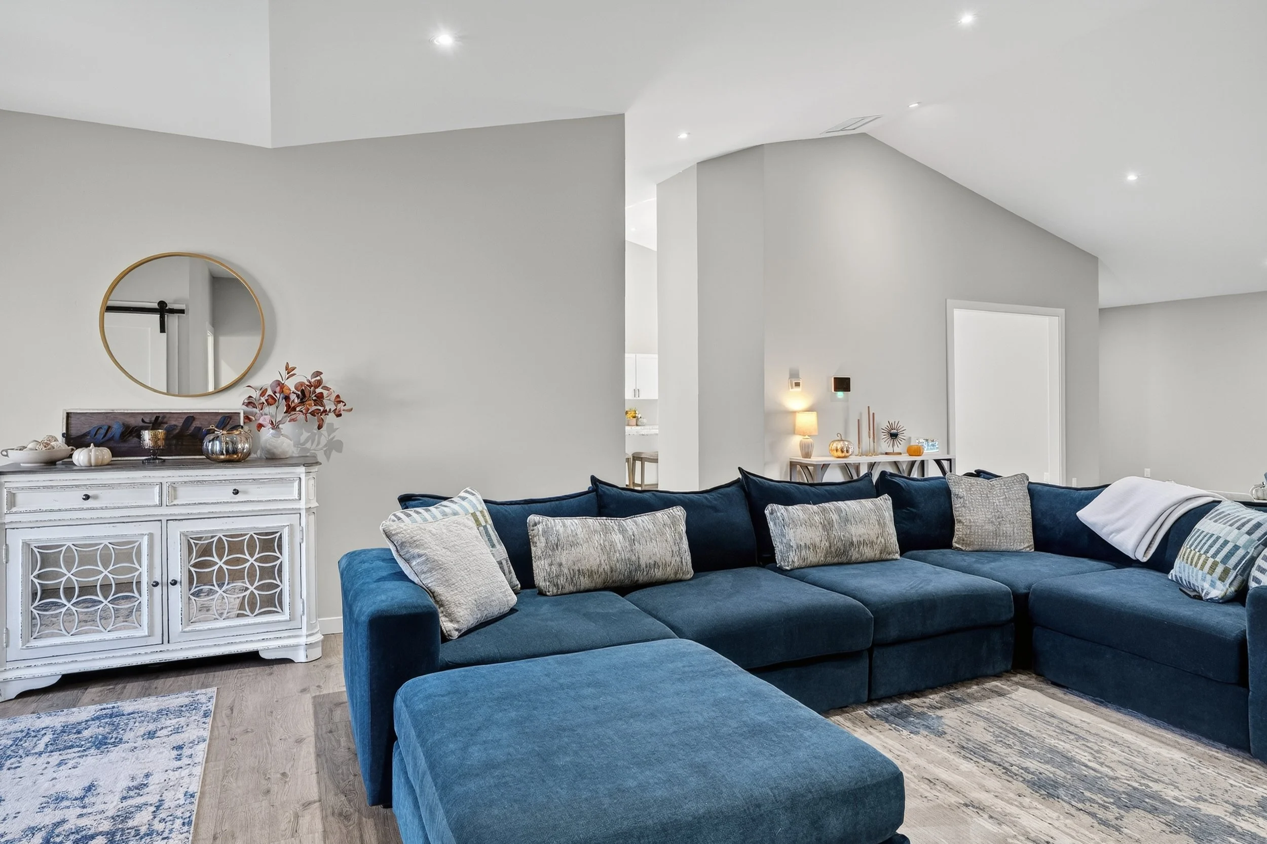 Living room with a large blue sectional sofa and decorative pillows, a white distressed sideboard with pumpkins and flowers, and a wall mirror.