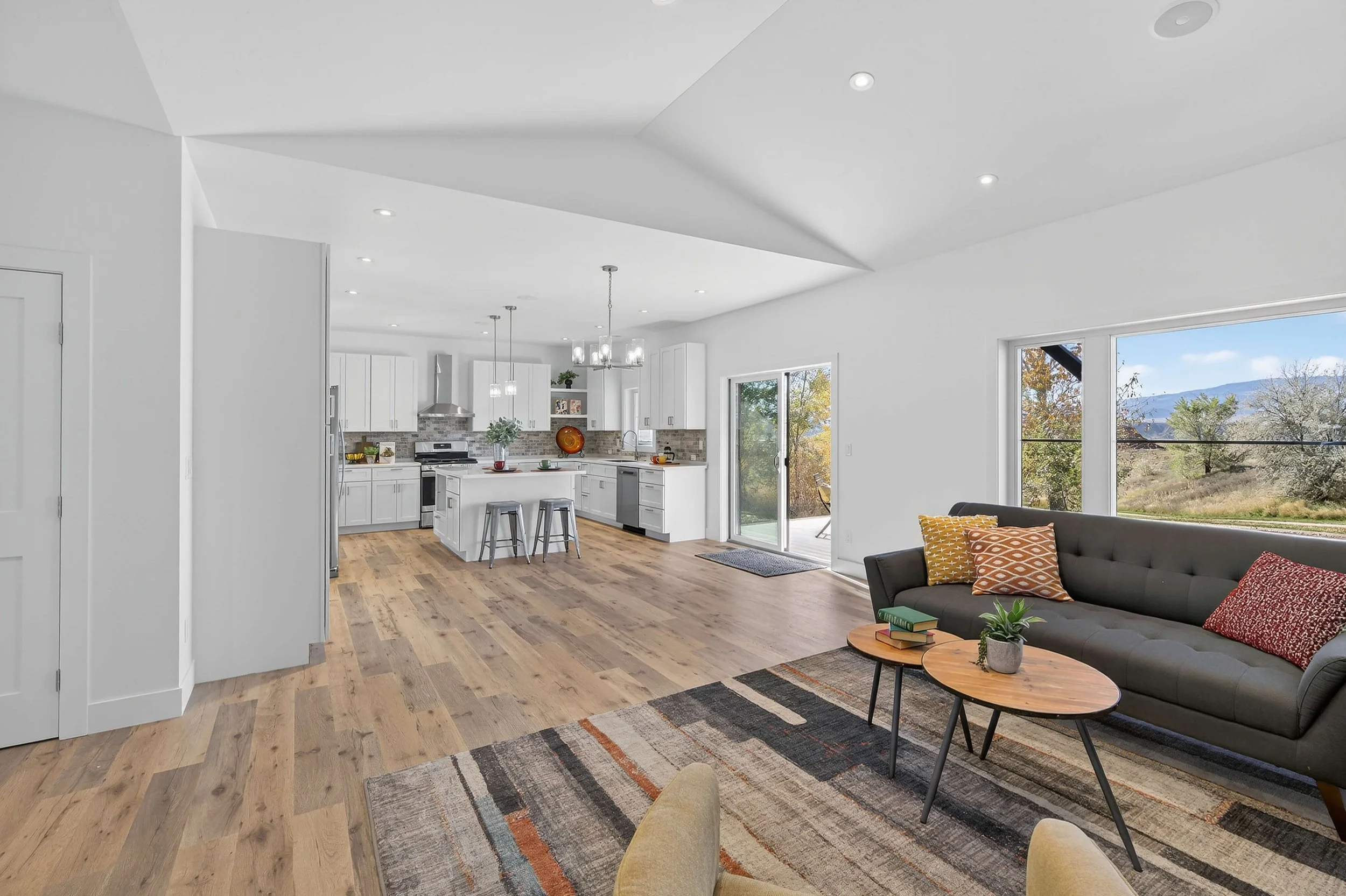 Open-concept living room and kitchen with white cabinets, stainless steel appliances, hardwood flooring, and large windows showing a scenic outdoor view.