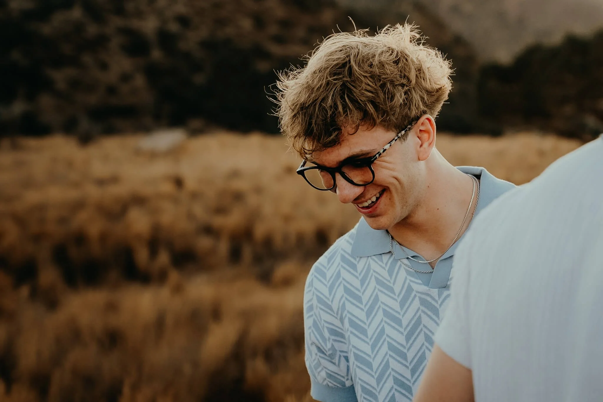 A young man with curly blond hair, glasses, and a silver necklace, smiling while looking down, outdoors in a natural setting with dry grass and rocky landscape.