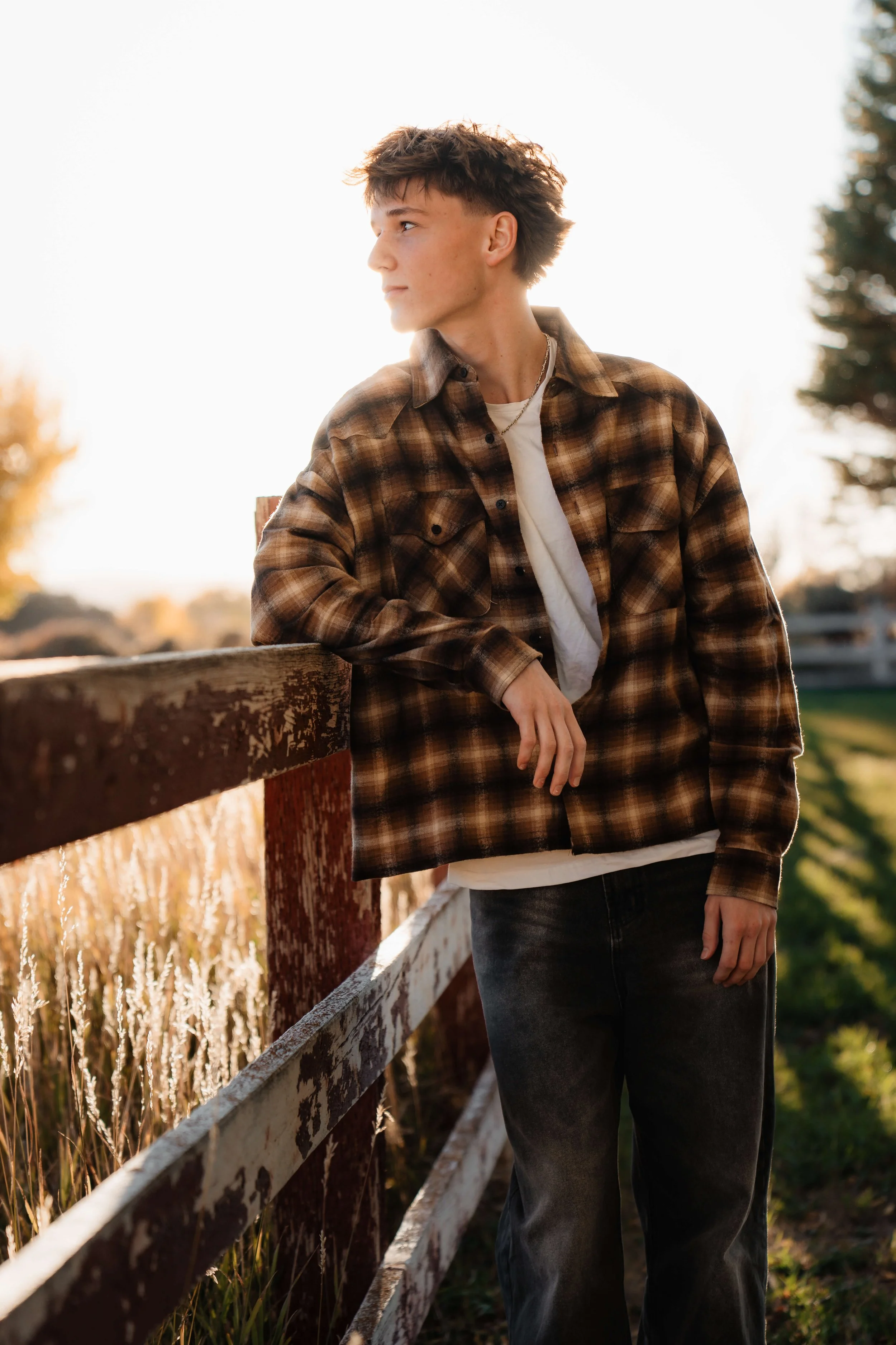 Young man wearing a plaid shirt and jeans, leaning on a wooden fence outdoors during sunset.