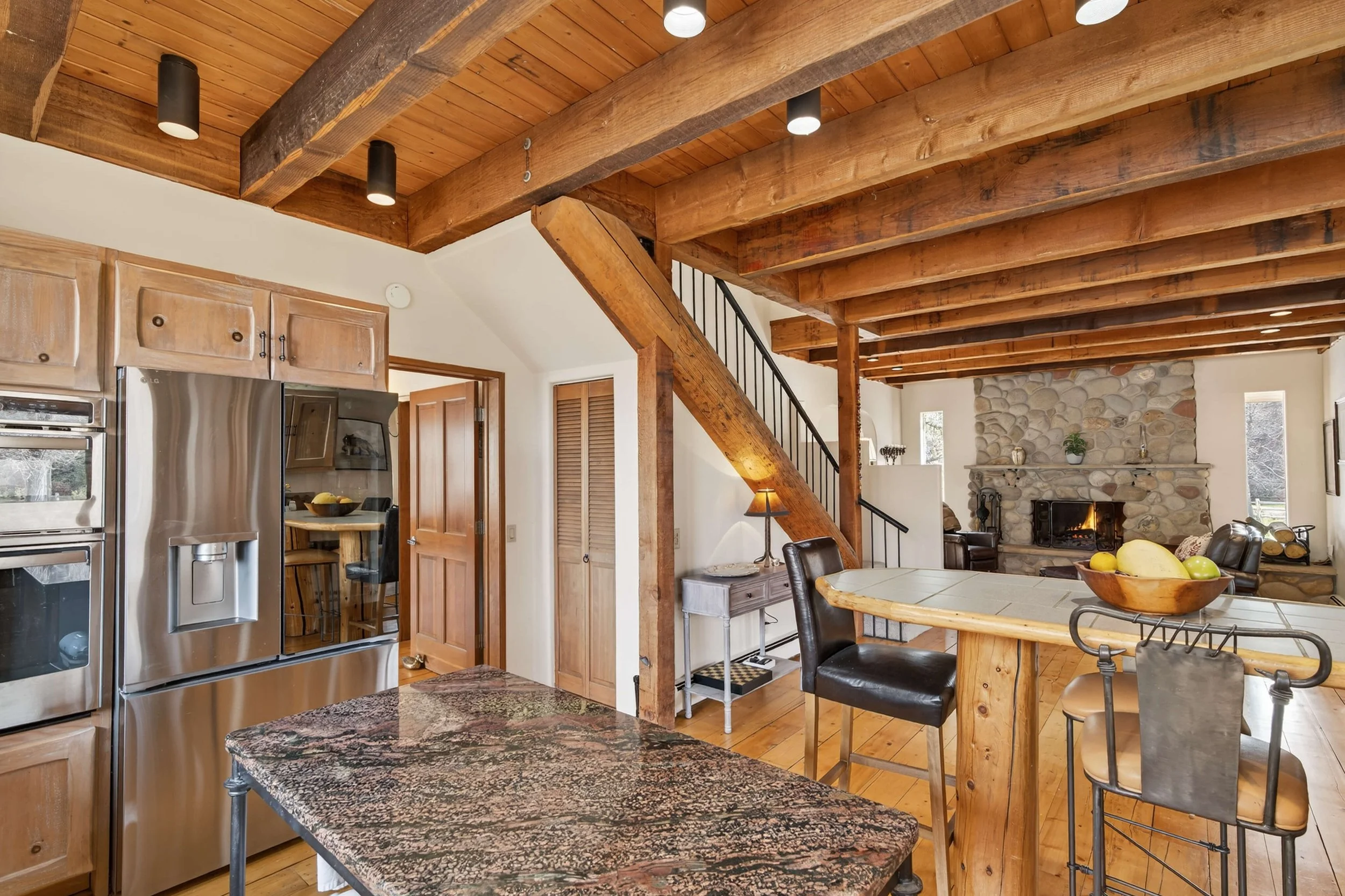 Open-concept kitchen and living room with wooden beams, stone fireplace, and stainless steel refrigerator.