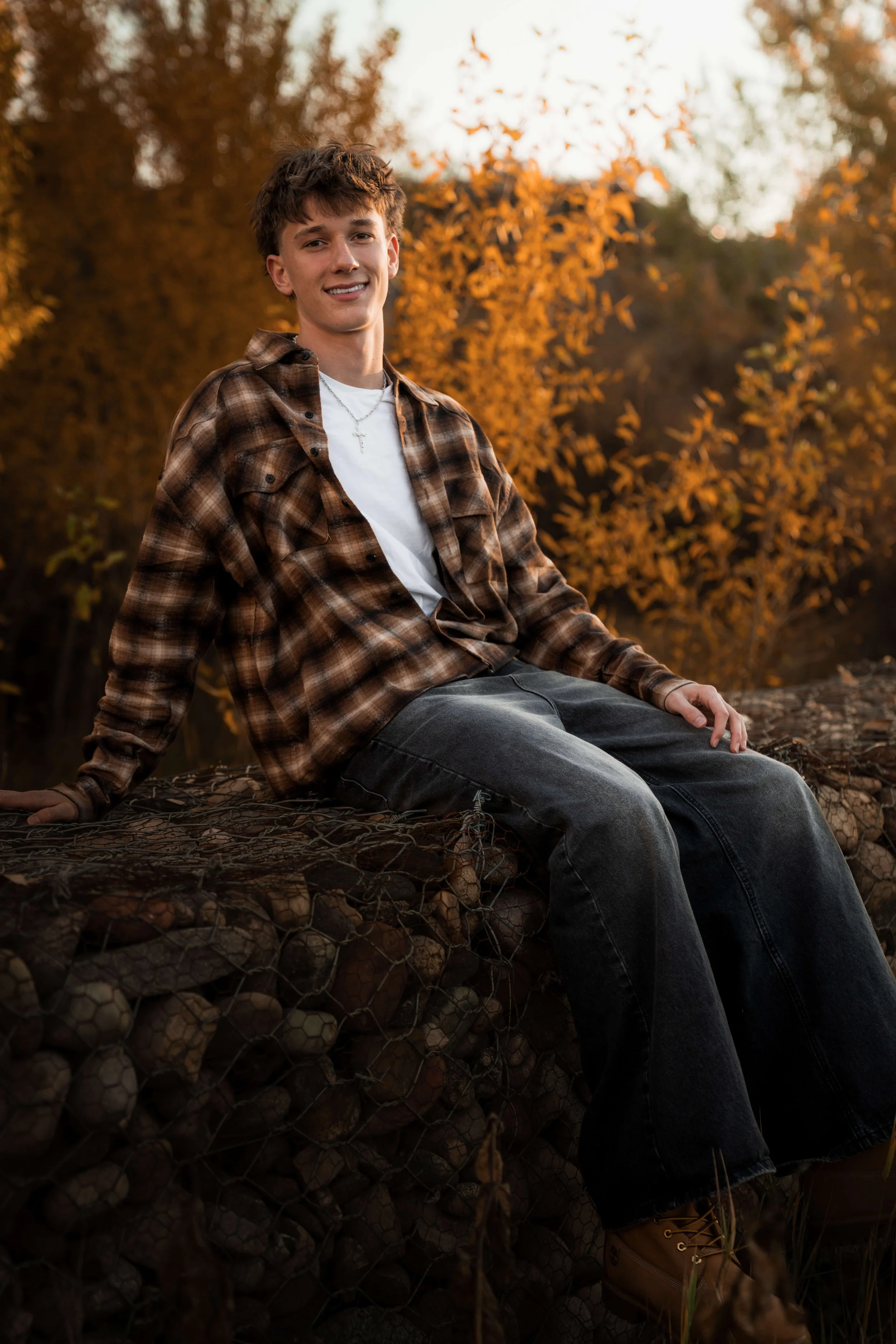 A young man sitting on a pile of logs outdoors during autumn, wearing a plaid shirt and jeans, with trees with orange leaves in the background.