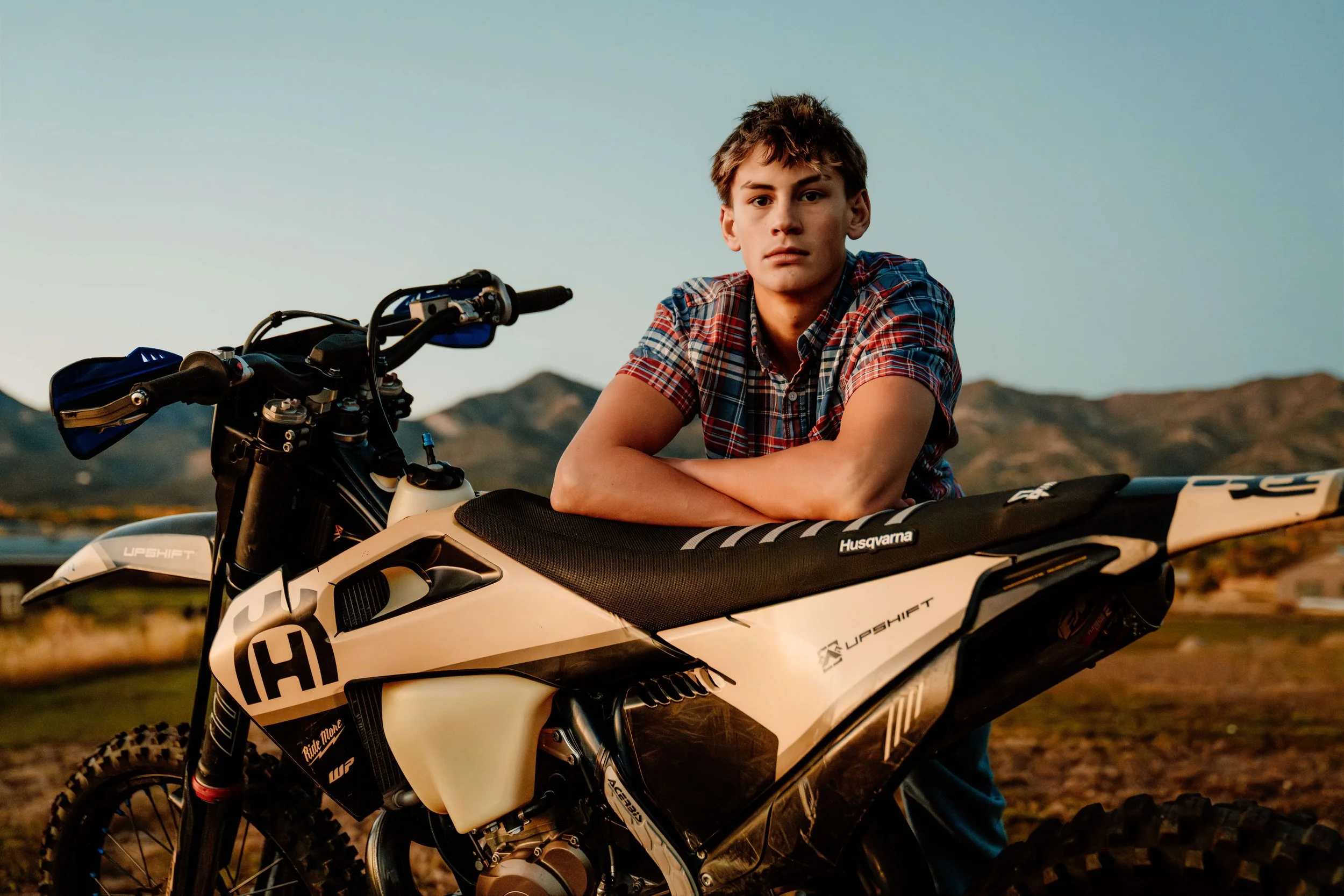 A young man in a plaid shirt leaning on a white Husqvarna dirt bike outdoors during sunset, with mountains in the background.