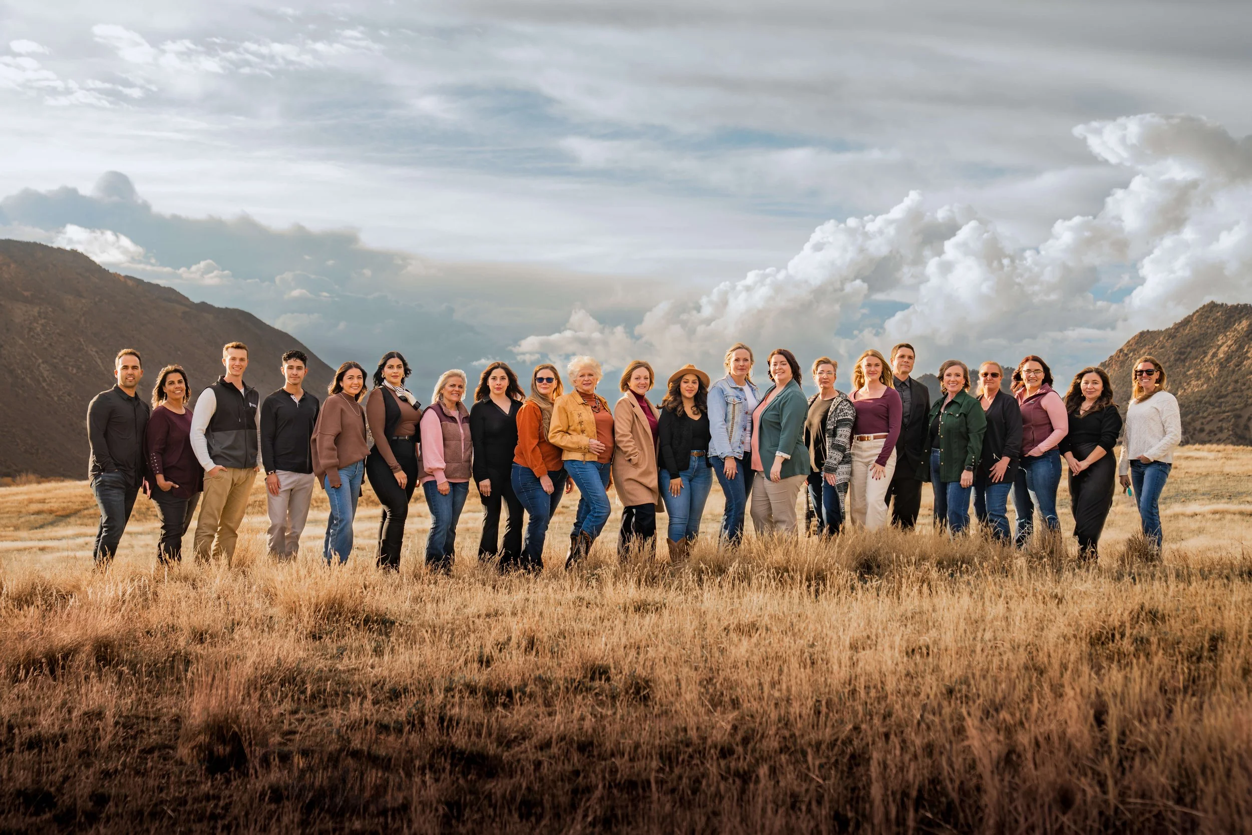 Group of diverse people standing in a grassy field with mountains and cloudy sky in the background.