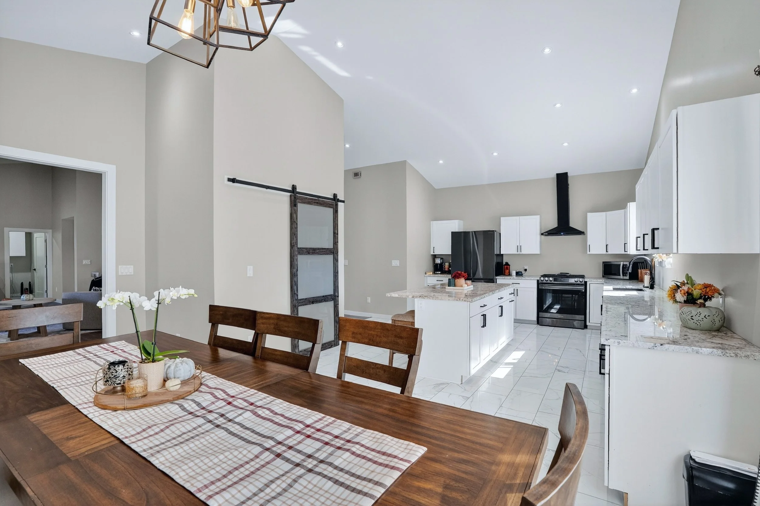 Modern kitchen with white cabinets, black appliances, and a marble island, adjacent to a dining area with a wooden table and plaid table runner, decorated with flowers and pumpkins.