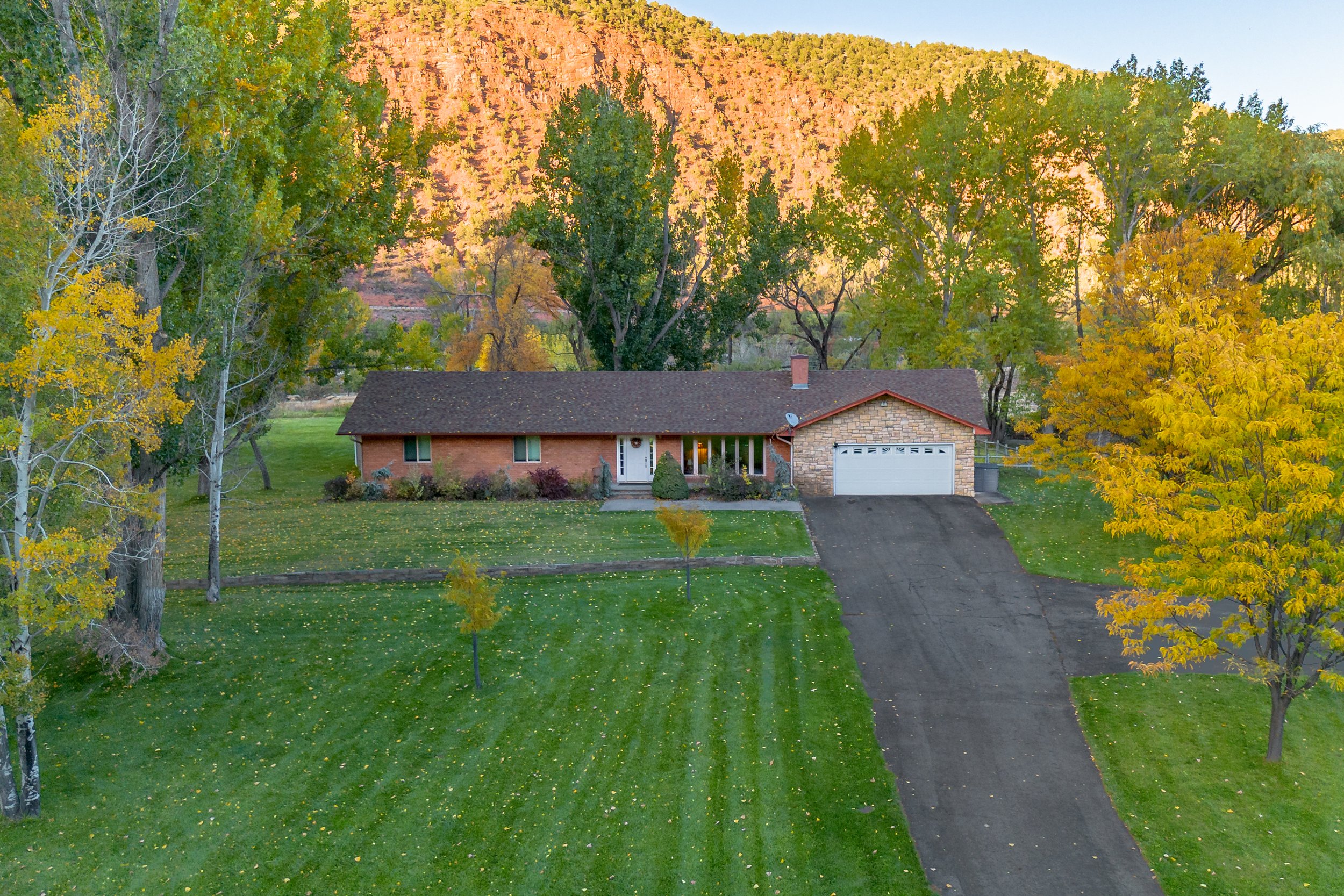 A single-story brick house with an attached garage, surrounded by a well-maintained green lawn with trees in fall colors, set against a backdrop of mountains under a clear sky.