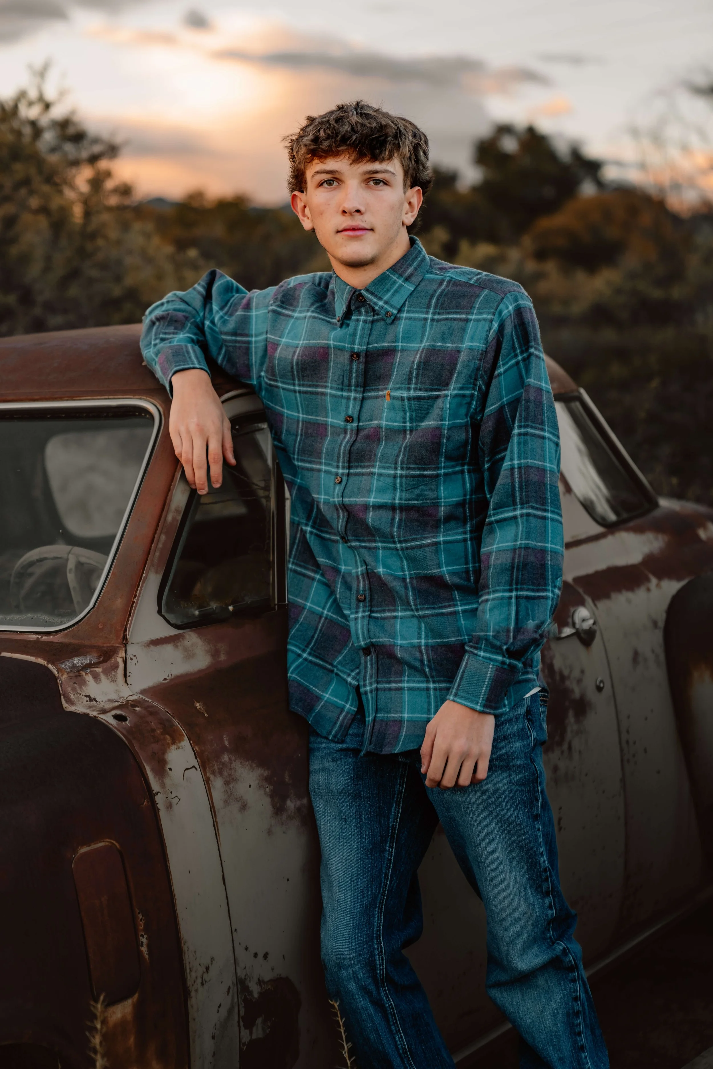 A young man with light brown, wavy hair and fair skin leaning against a rusty, vintage car during sunset, wearing a blue plaid shirt and jeans.