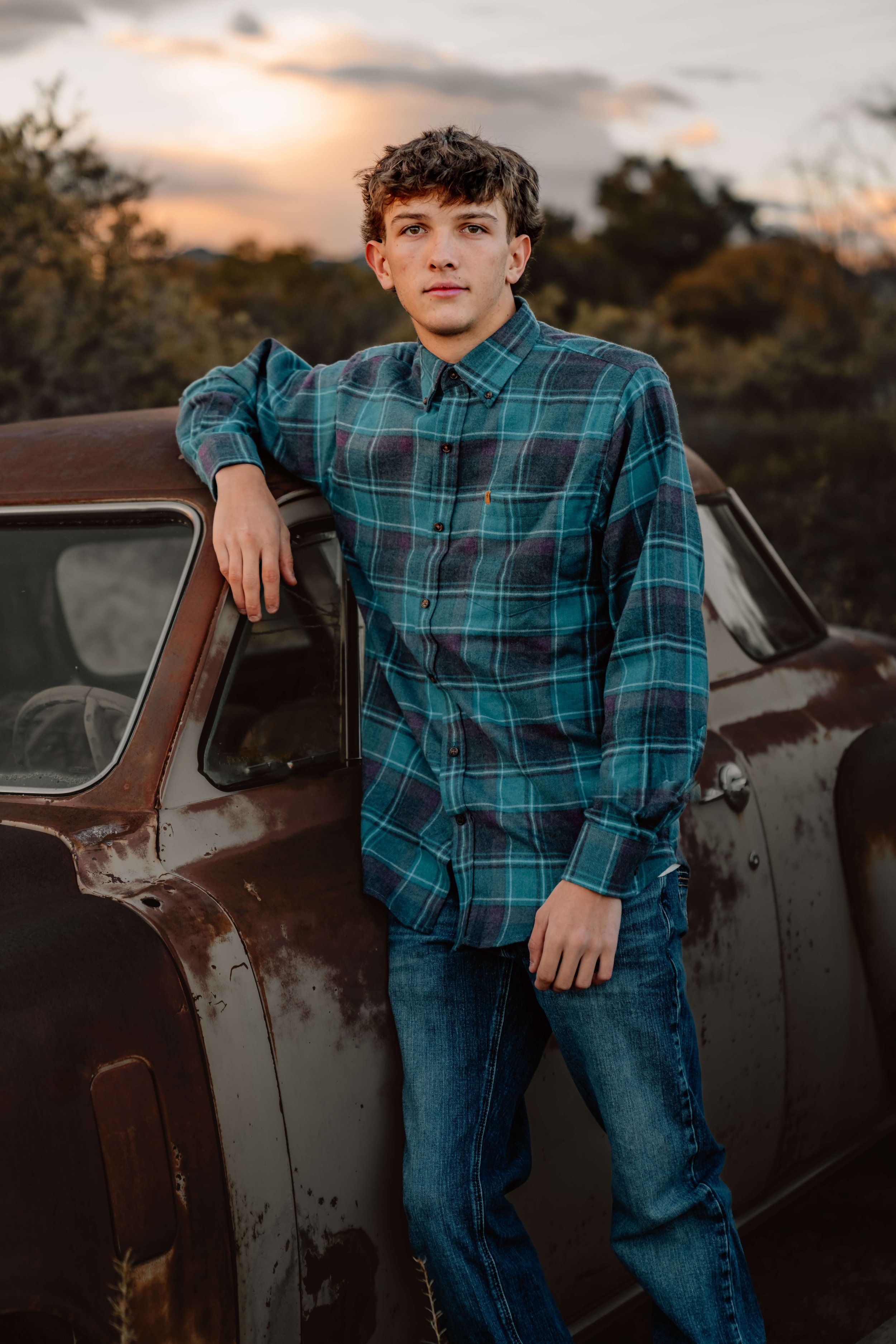A young man with light brown, wavy hair and fair skin leaning against a rusty, vintage car during sunset, wearing a blue plaid shirt and jeans.