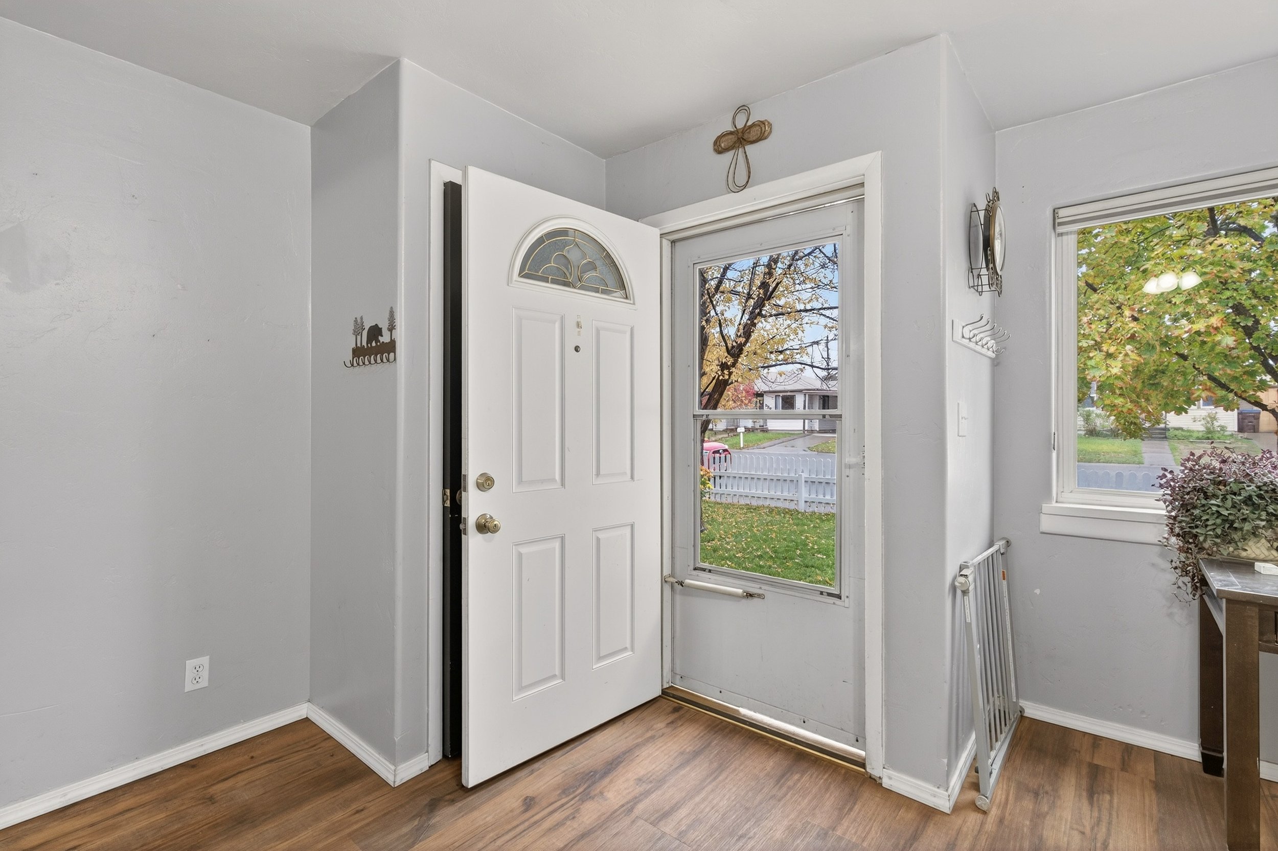 Interior view of a home entryway with a white front door, a window to the right showing autumn trees, and a small table with a potted plant. The floor is hardwood, and the walls are painted light gray.