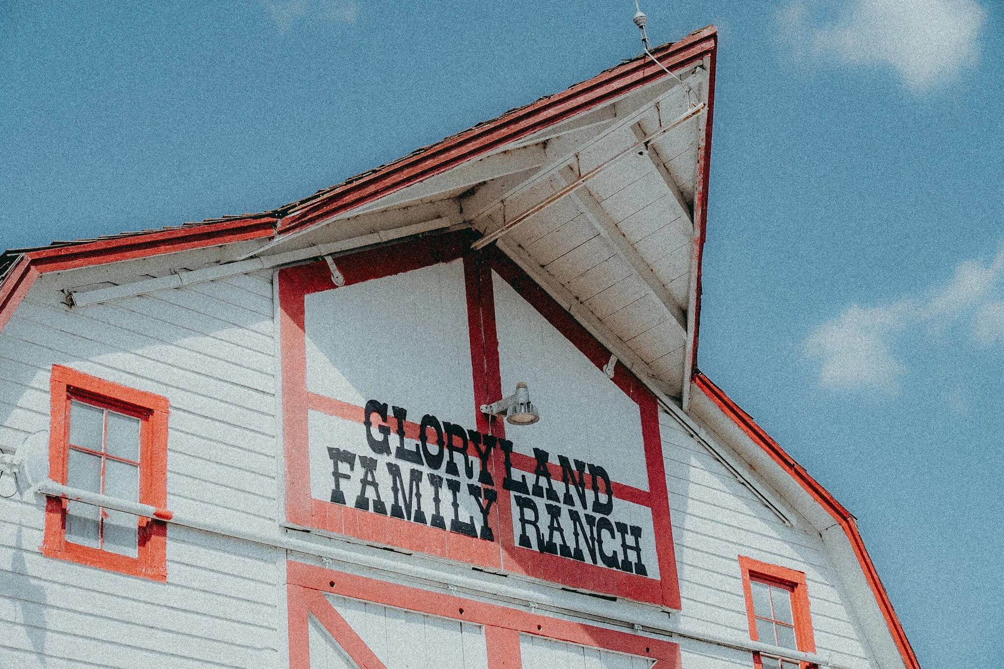 Close-up of a white barn with red trim, with a sign that reads 'Glory Land Family Ranch' on the front. The sky is blue with a few clouds.