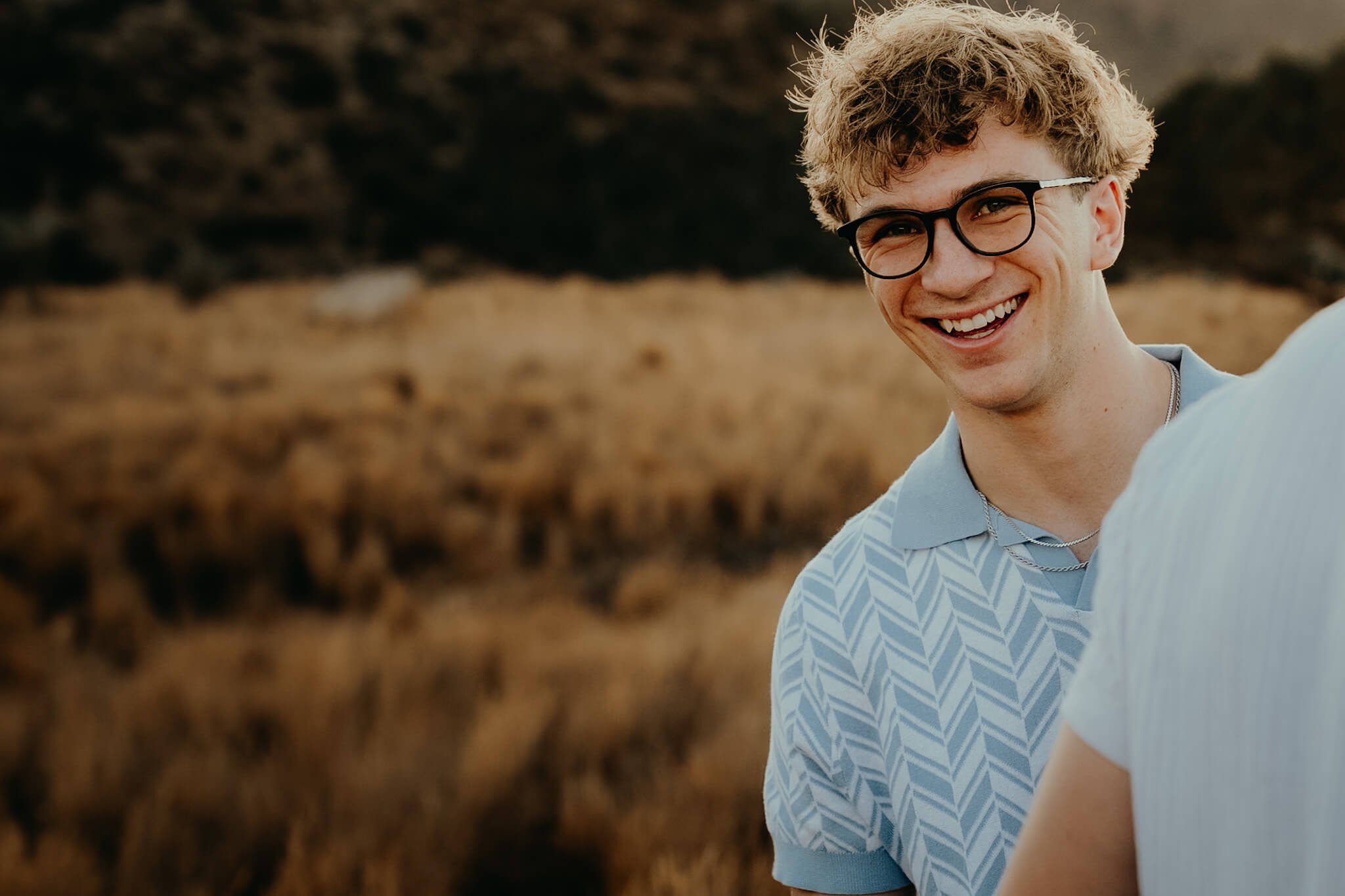Close-up of a young man with curly blond hair, black glasses, and a light blue patterned shirt smiling outdoors with a blurred natural landscape in the background.