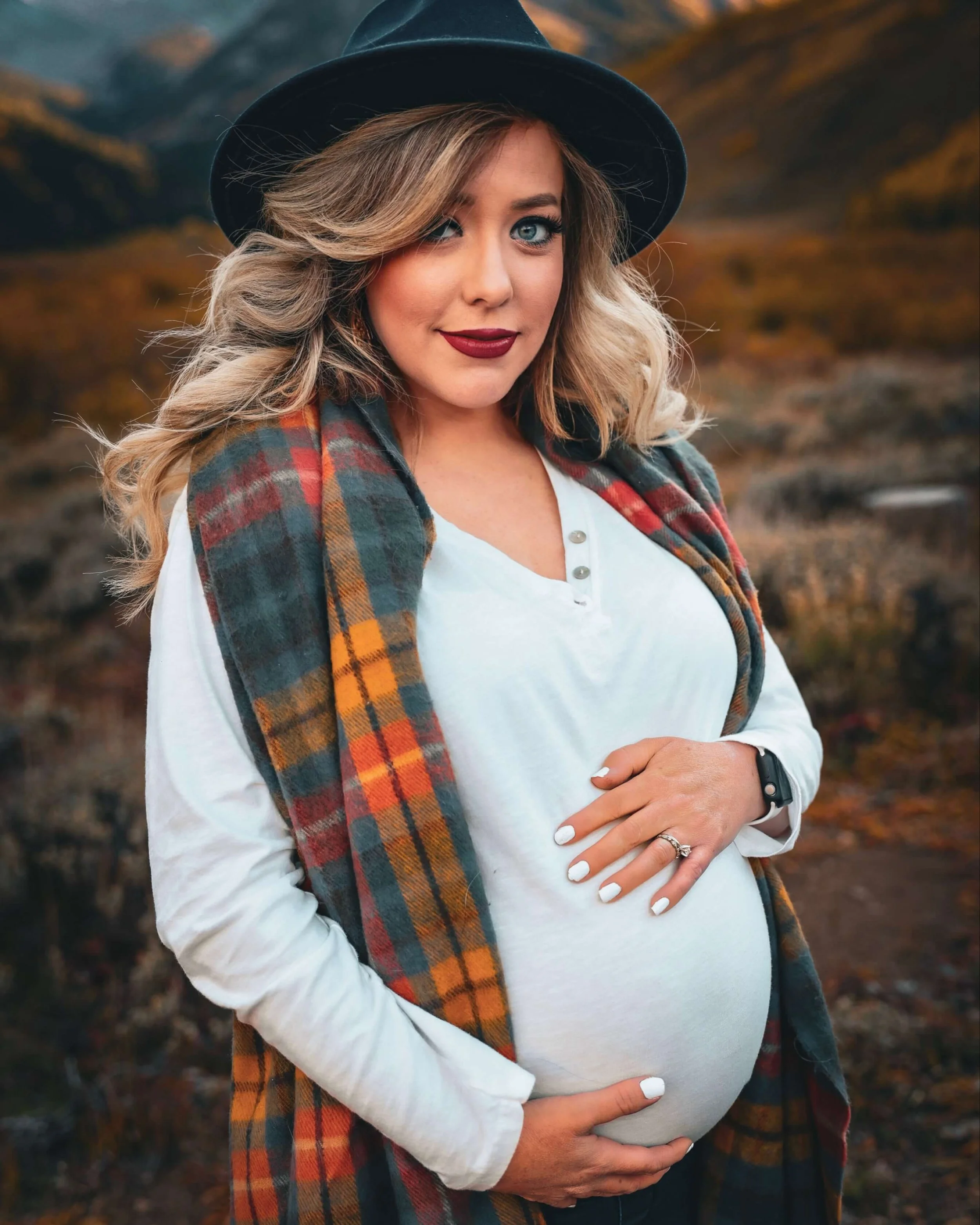 A woman with blonde, wavy hair, blue eyes, and fair skin, wearing a black hat, white shirt, and a multicolored plaid scarf, stands outdoors in a mountainous area during fall, gently cradling her pregnant belly.