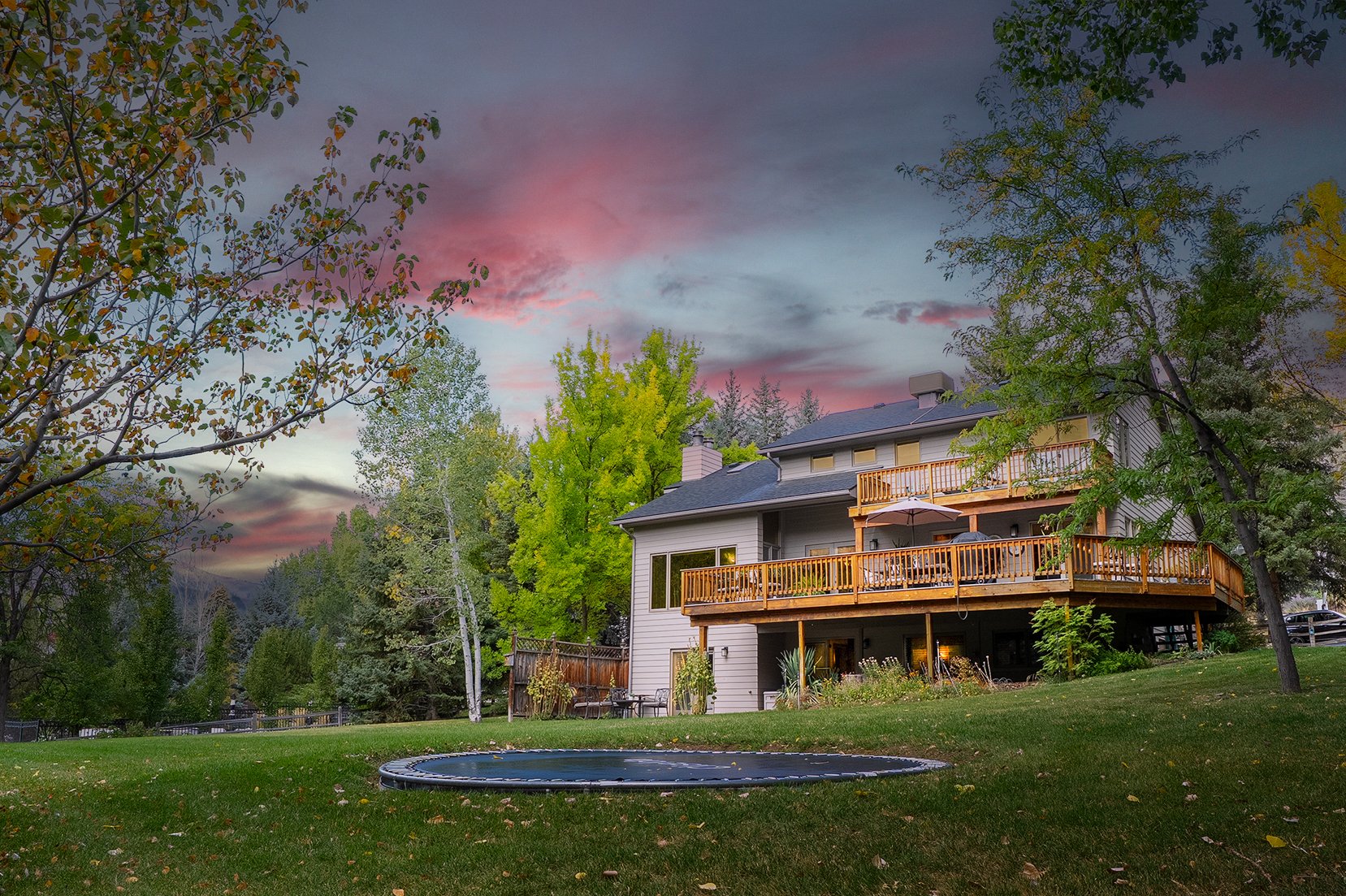 A two-story house with a large wooden deck in a green yard with trees and a small round trampoline, sunset sky with pink and purple clouds.