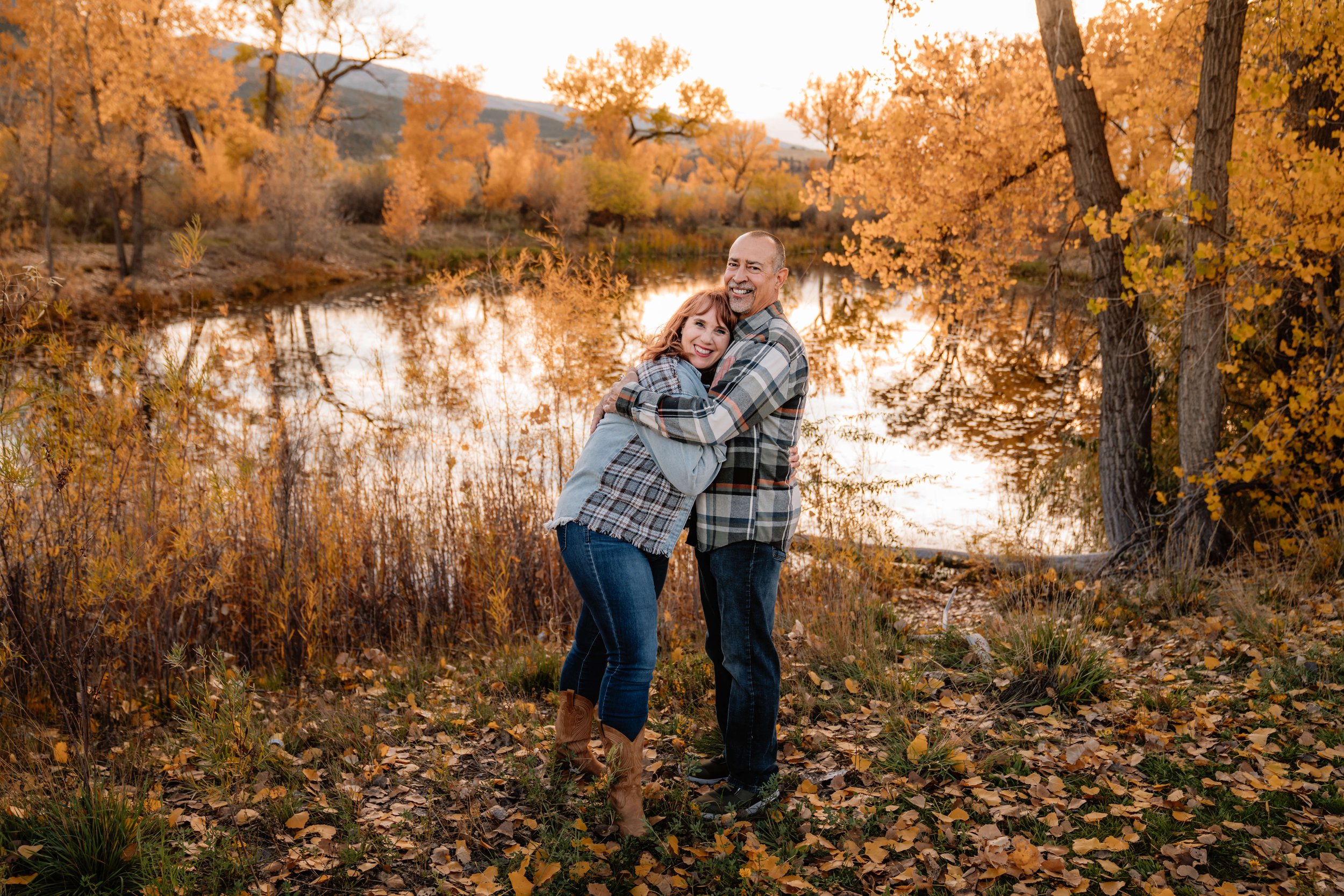 A smiling couple embracing outdoors during autumn near a lake, surrounded by trees with orange and yellow leaves, with a mountain in the background.