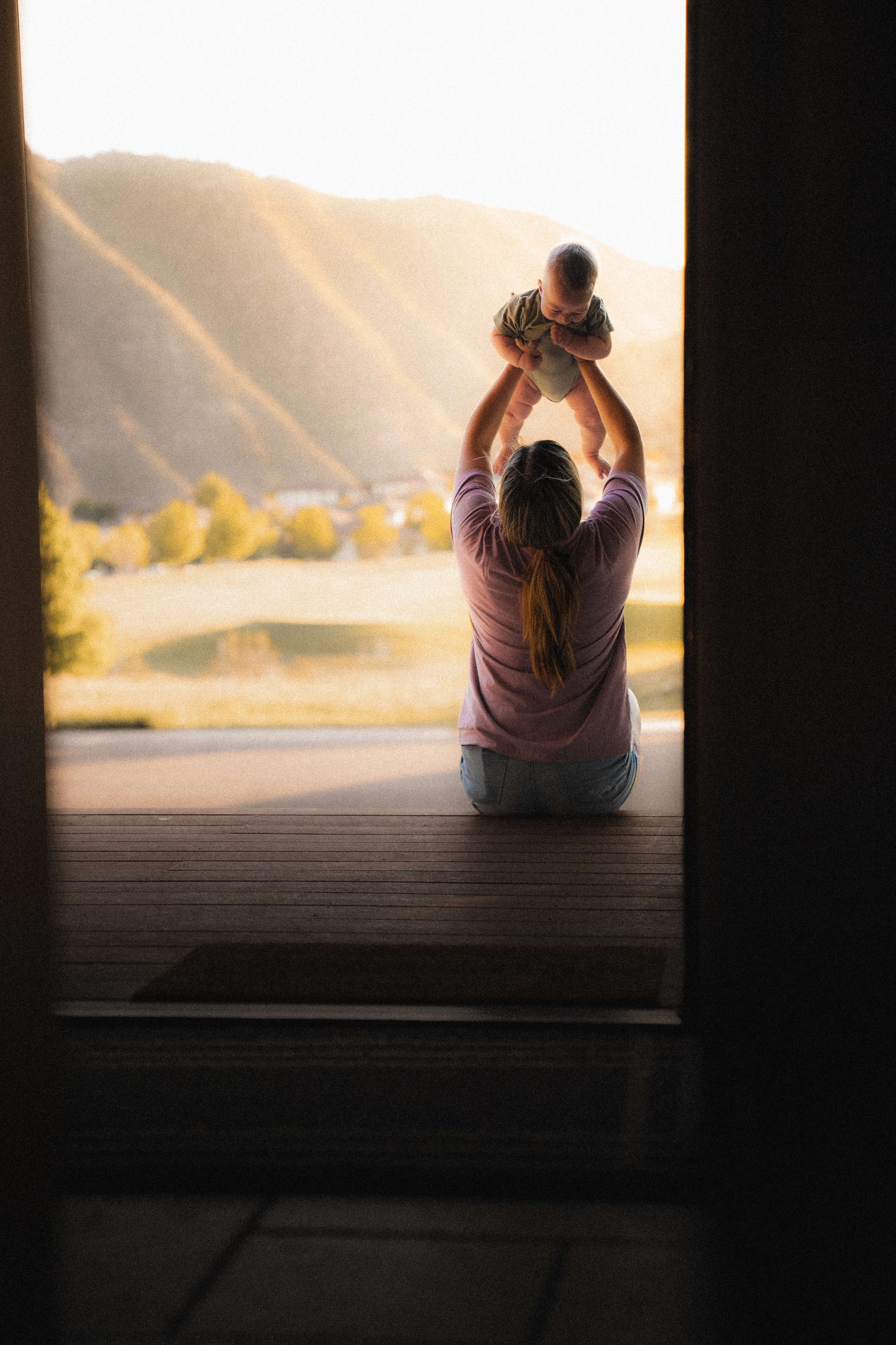 A woman sitting on a porch holding a baby up in the air with mountains in the background during sunset.