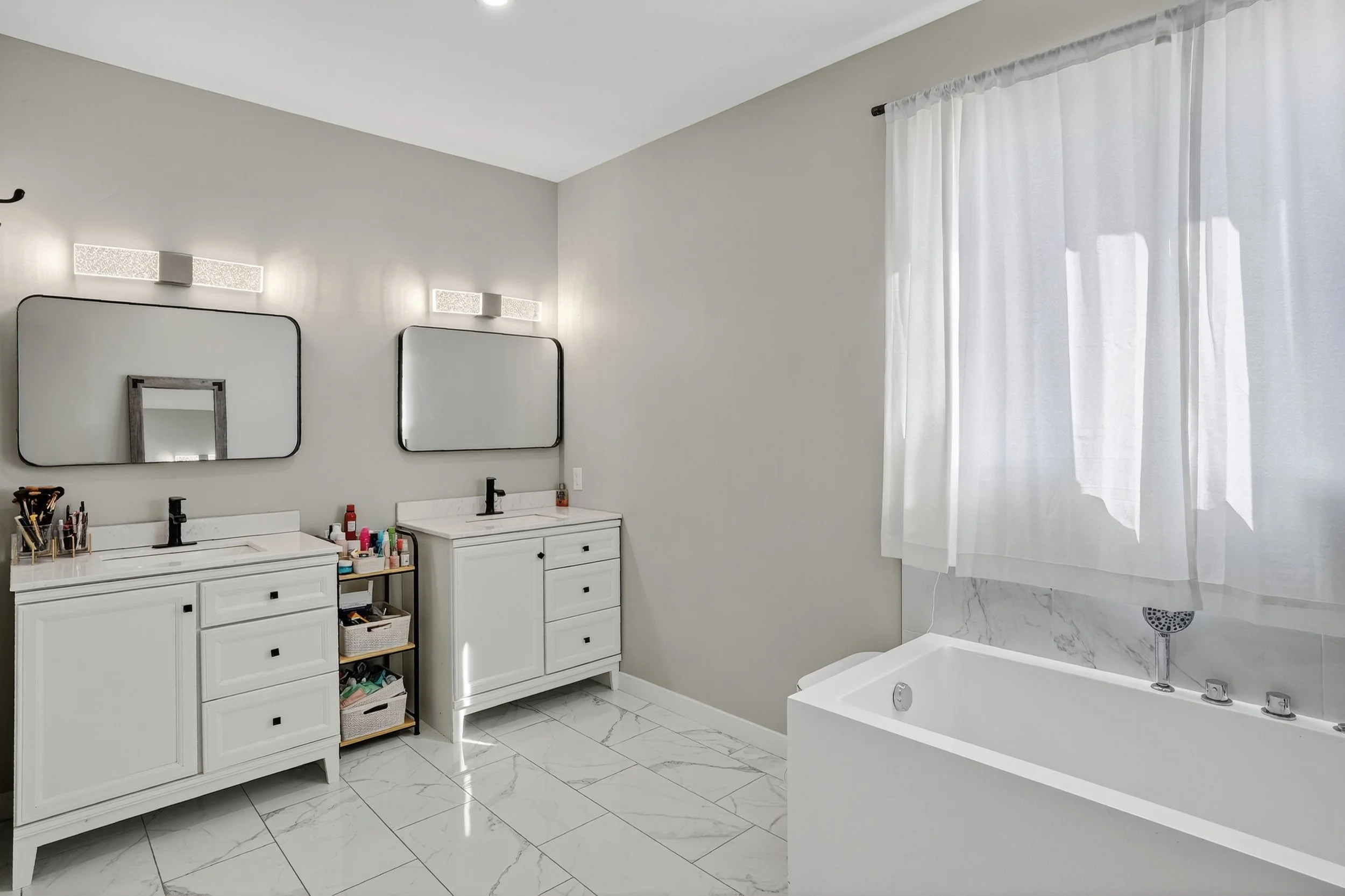 A bright bathroom with two white vanities with black fixtures, large rectangular mirrors, and light above. There is a small black shelving unit between the vanities holding toiletries. A white bathtub with a marble backsplash and a window with white 