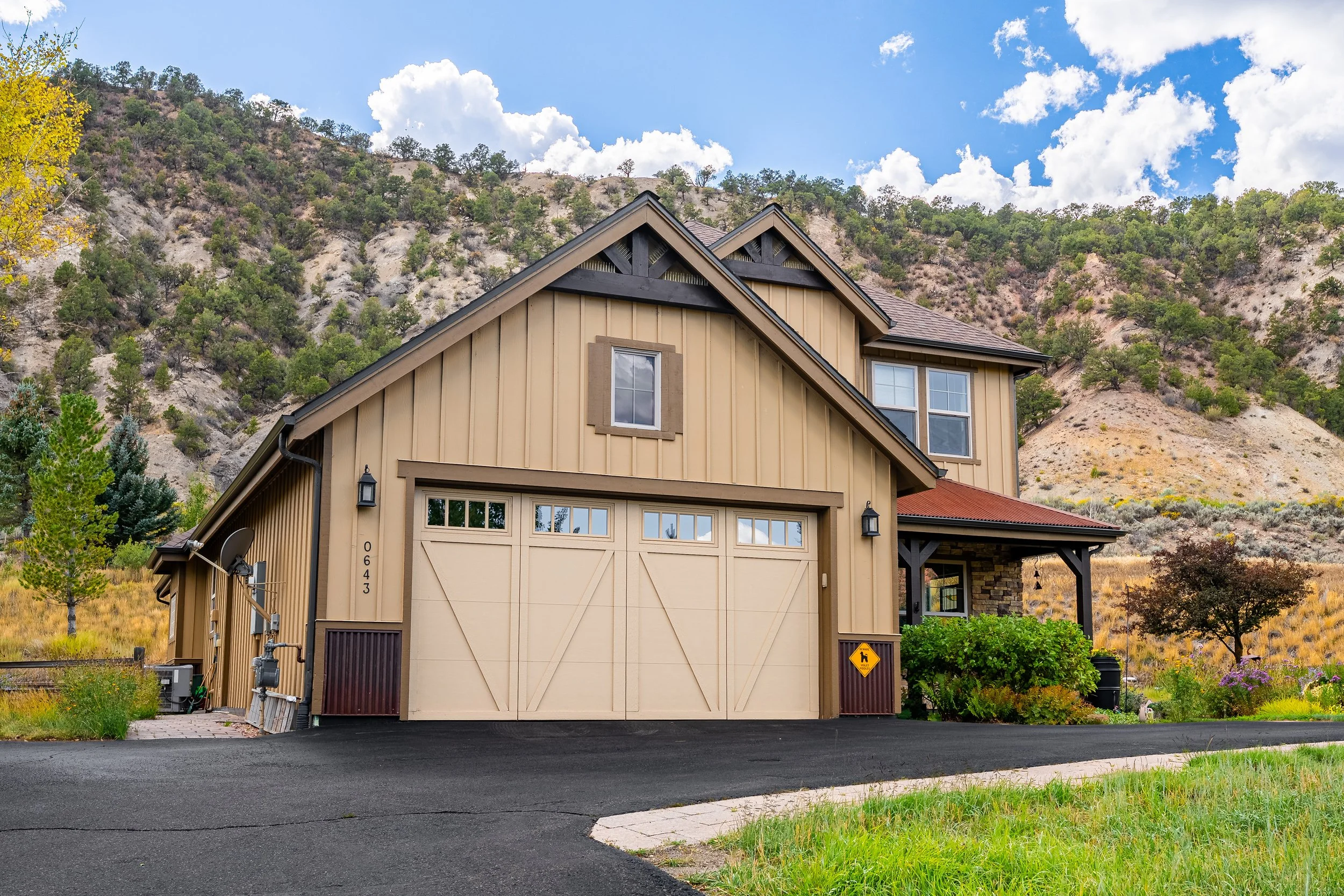 A modern two-story house with beige vertical siding and a multi-gabled roof set against a hilly, green landscape under a blue sky with clouds.