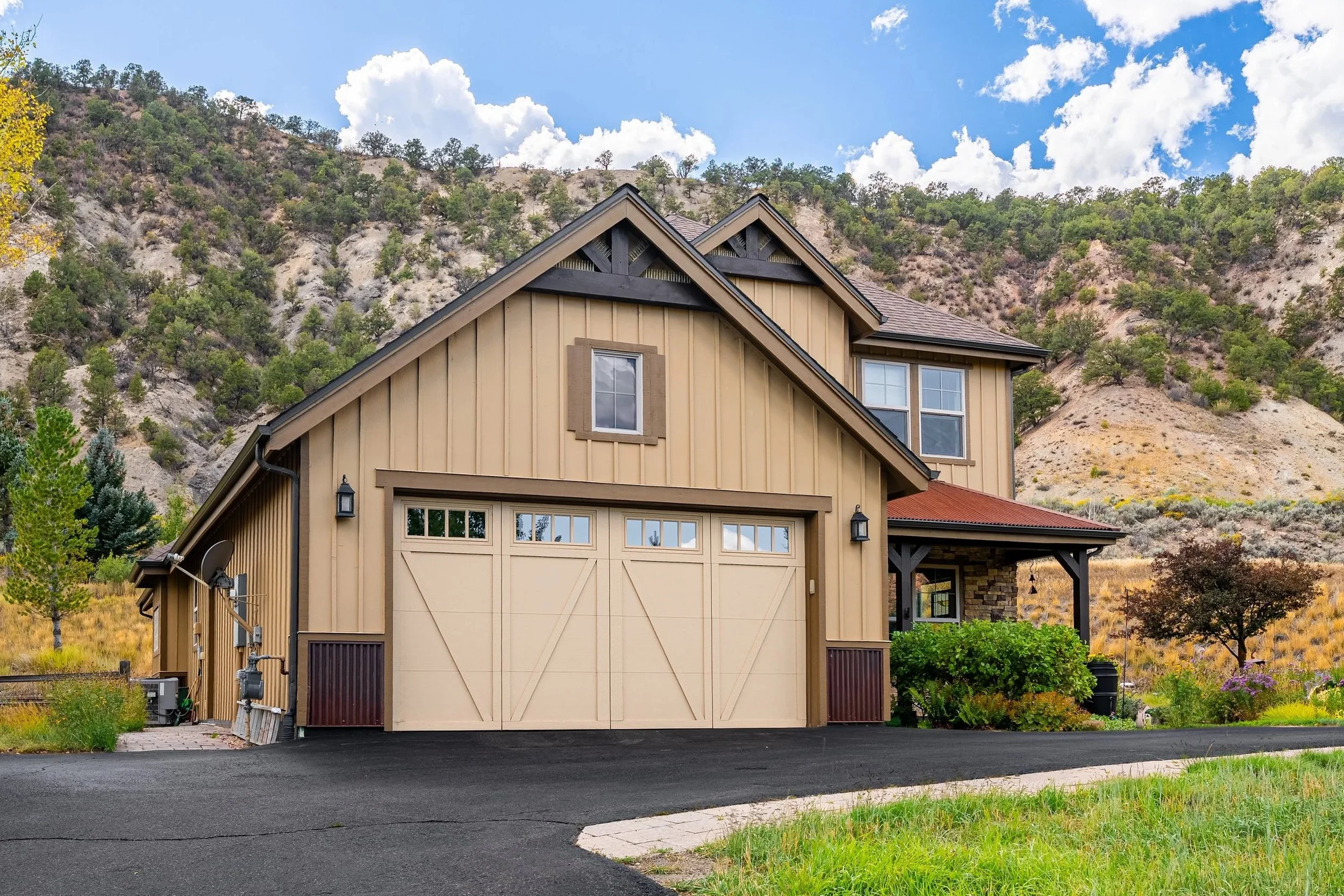 A two-story house with a beige garage door, surrounded by green plants, set against a scenic hillside with trees and shrubs under a partly cloudy sky.