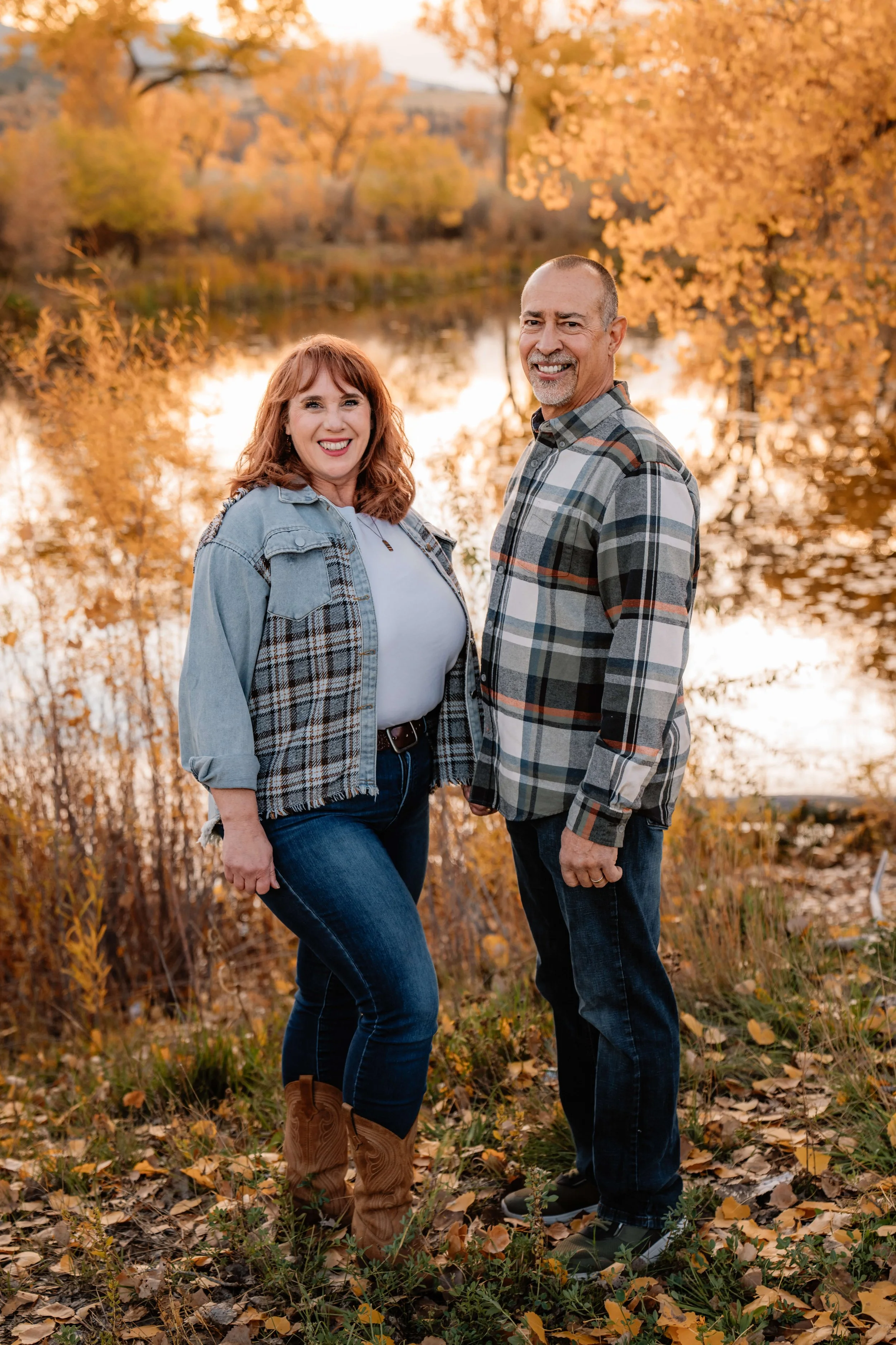 A smiling couple standing outdoors near a body of water during autumn, surrounded by fall foliage, with a river and trees with orange and yellow leaves in the background.