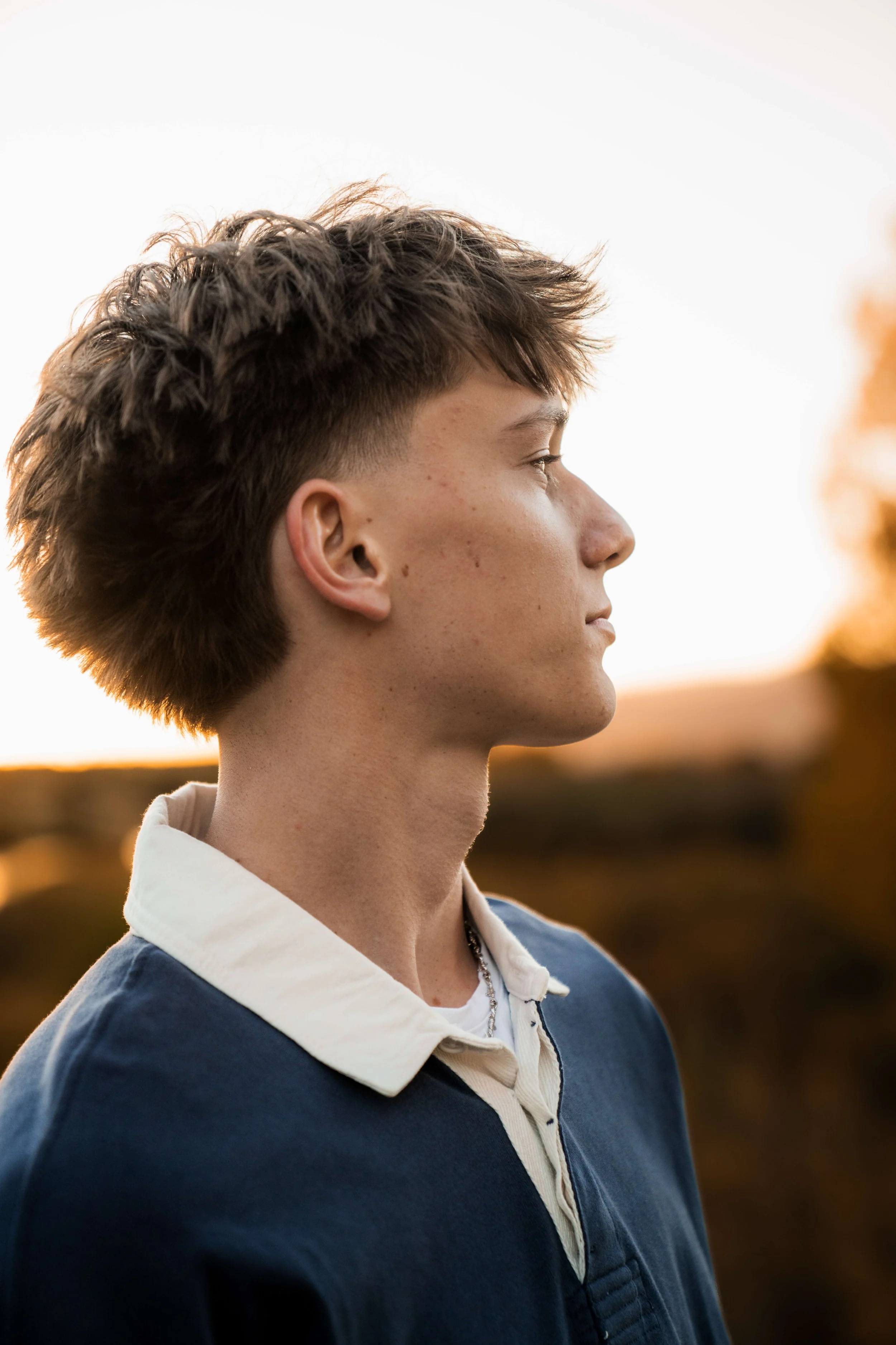 Profile of a young man with short, textured brown hair, wearing a blue jacket with a white collar, standing outdoors during sunset.