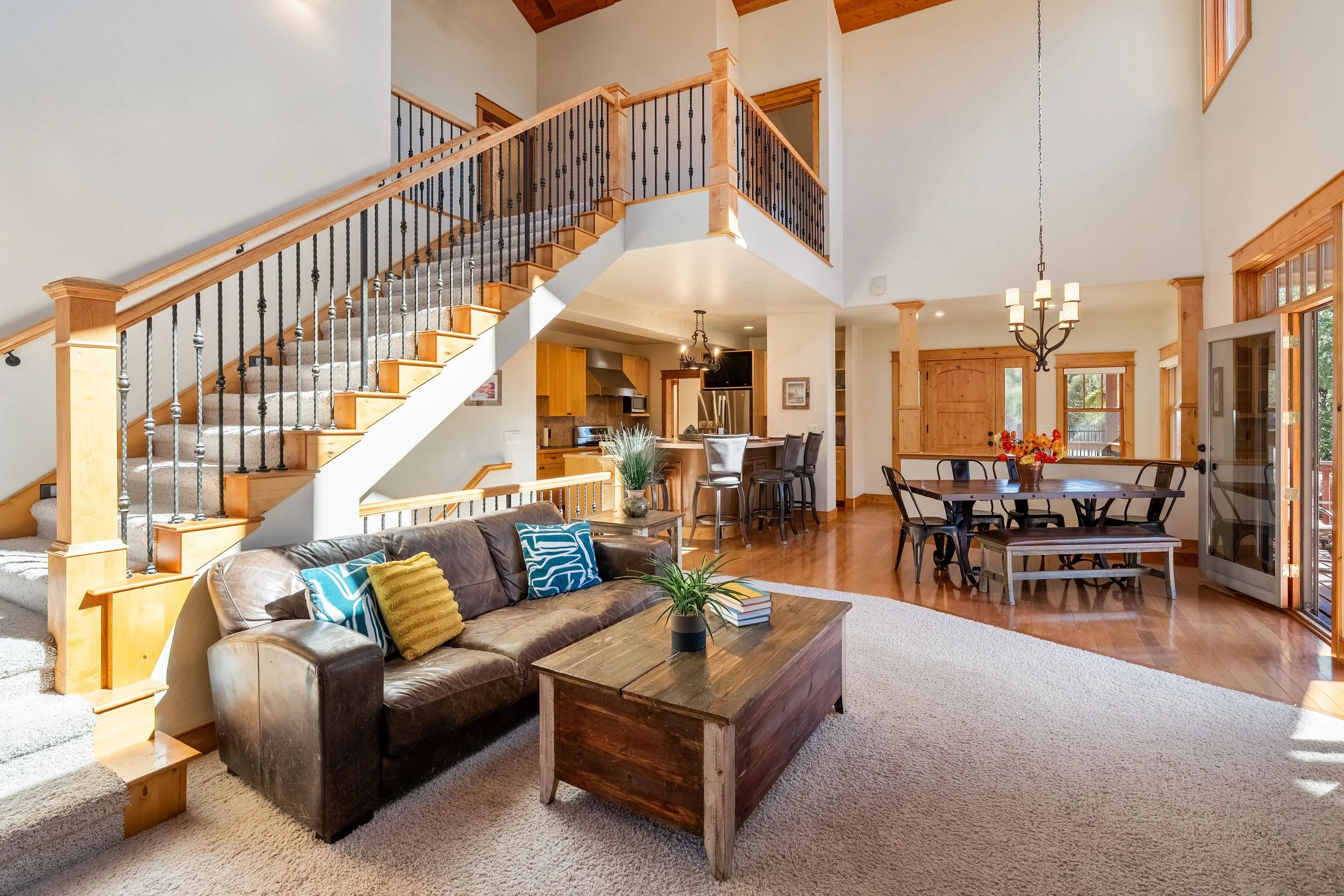 Living area with a leather sofa, wooden coffee table, dining table with chairs, open kitchen, staircase, and large windows letting in natural sunlight.