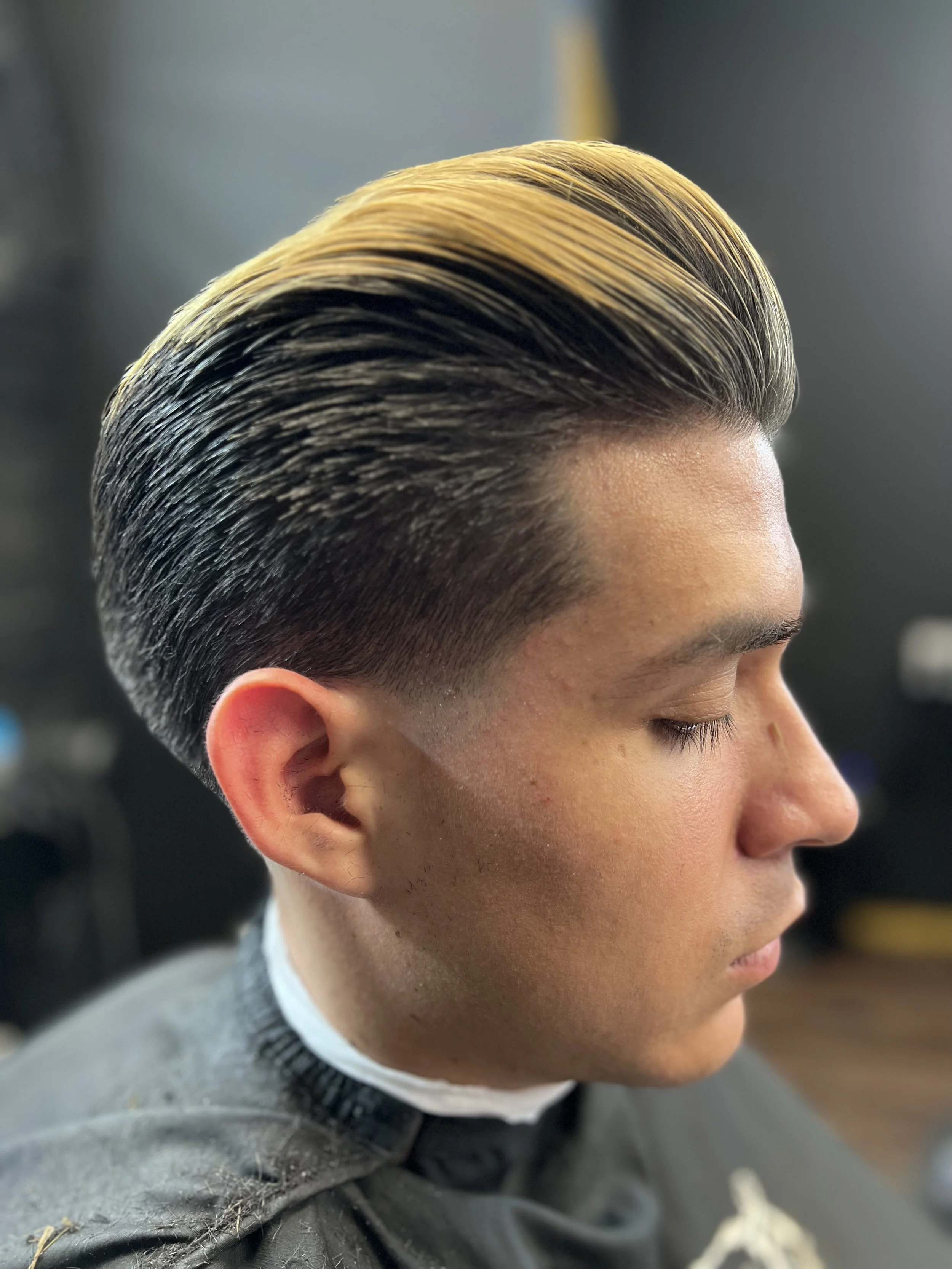 Side view of a young man with a slicked-back hairstyle featuring dark roots and blonde highlights on top, in a barbershop setting.