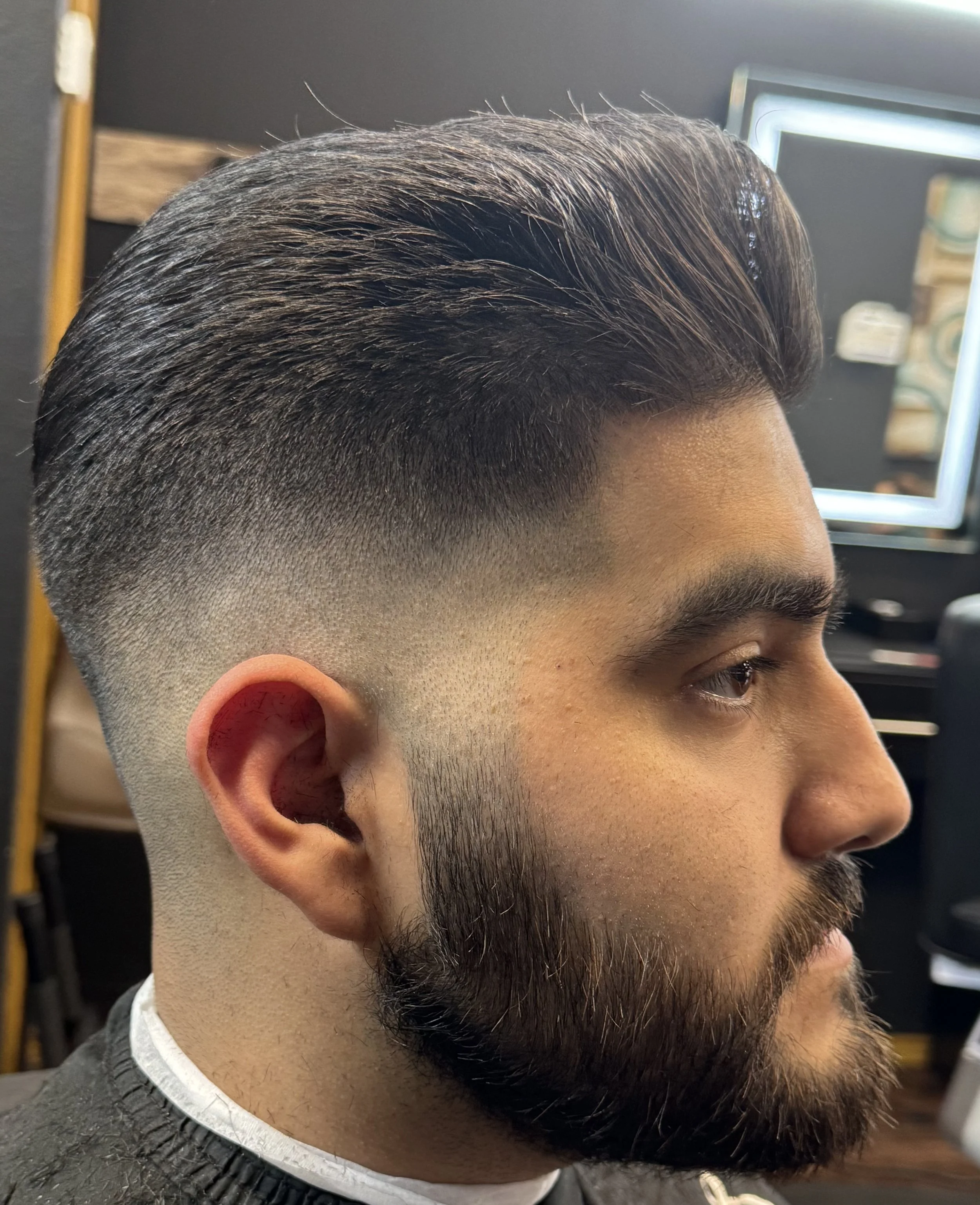 Side profile of a man with a modern fade haircut, featuring short sides and longer hair on top, with a full beard.