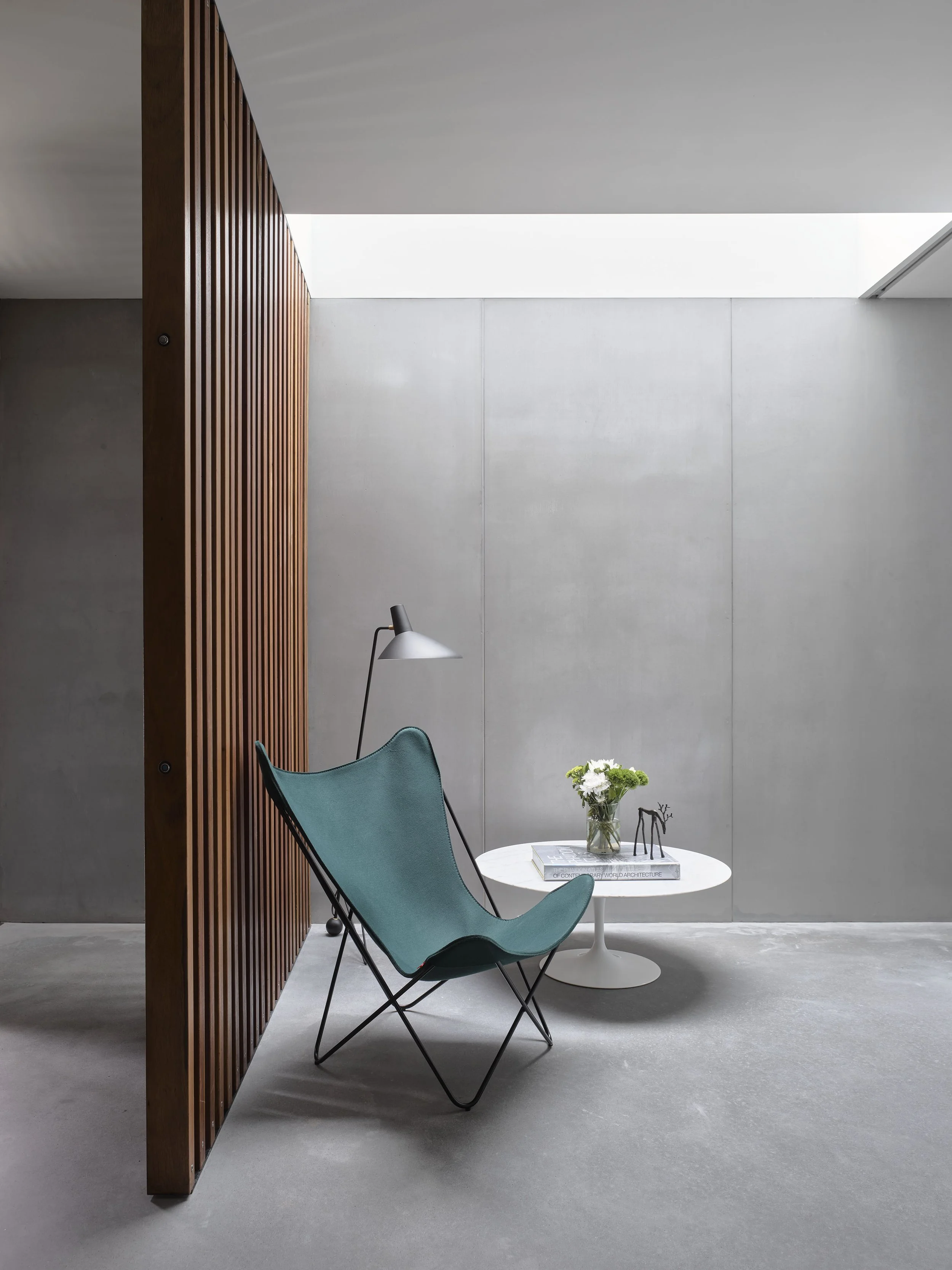 Modern minimalist interior with a teal butterfly chair, a white round coffee table with a flower vase, and a black floor lamp, in a space with concrete walls and a ceiling with natural light.