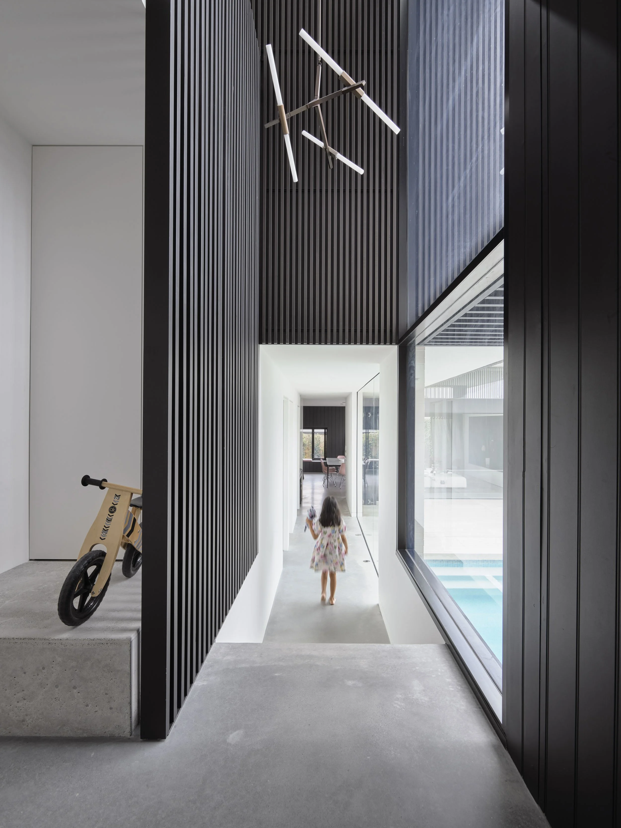 Interior view of a modern home with black and white walls, a wooden balance bike, and a girl walking down a hallway toward a bright room with a pool outside.