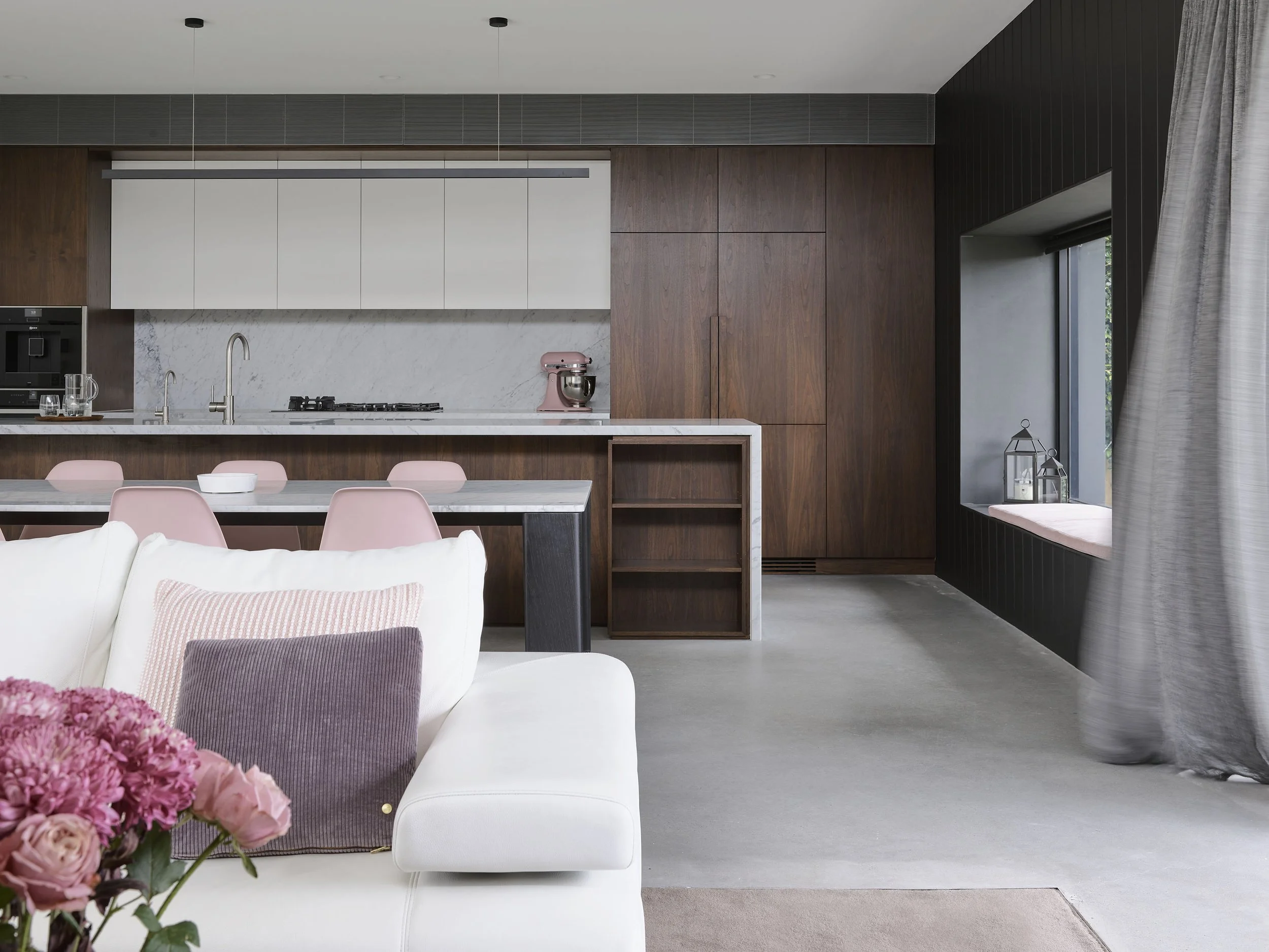 Modern kitchen with wooden cabinets, marble backsplash, pink kitchen appliances, and a dining area with pink chairs. In the foreground, part of a white sofa with pink and purple pillows and a bouquet of pink flowers.