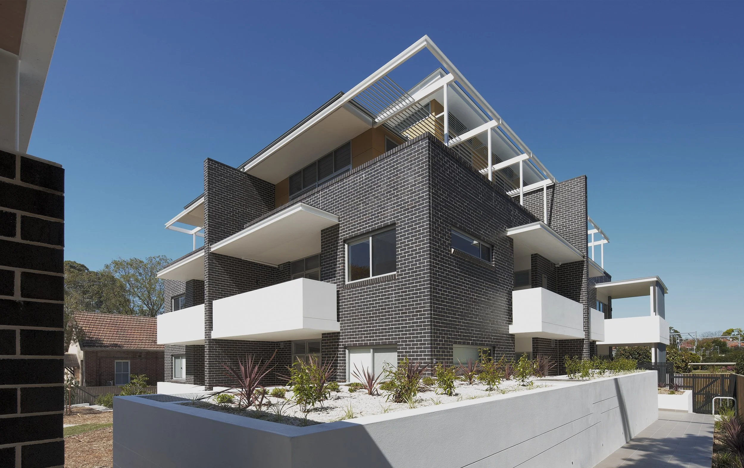 Modern multi-story apartment building with black brick exterior, white balconies, and rooftop terraces under a clear blue sky.