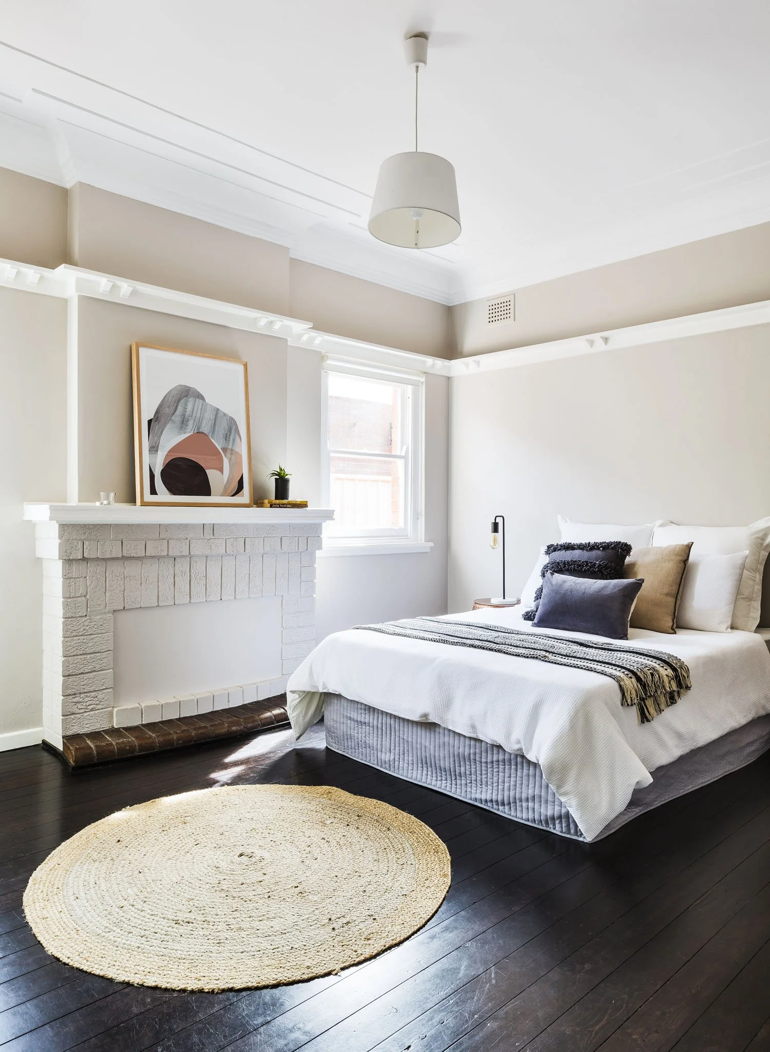 A bedroom with white walls, a white brick fireplace with a framed abstract art piece and small potted plant on top, a window letting in natural light, a white bed with multiple pillows and a striped blanket, a circular woven rug on dark hardwood floor, and a pendant light hanging from the ceiling.