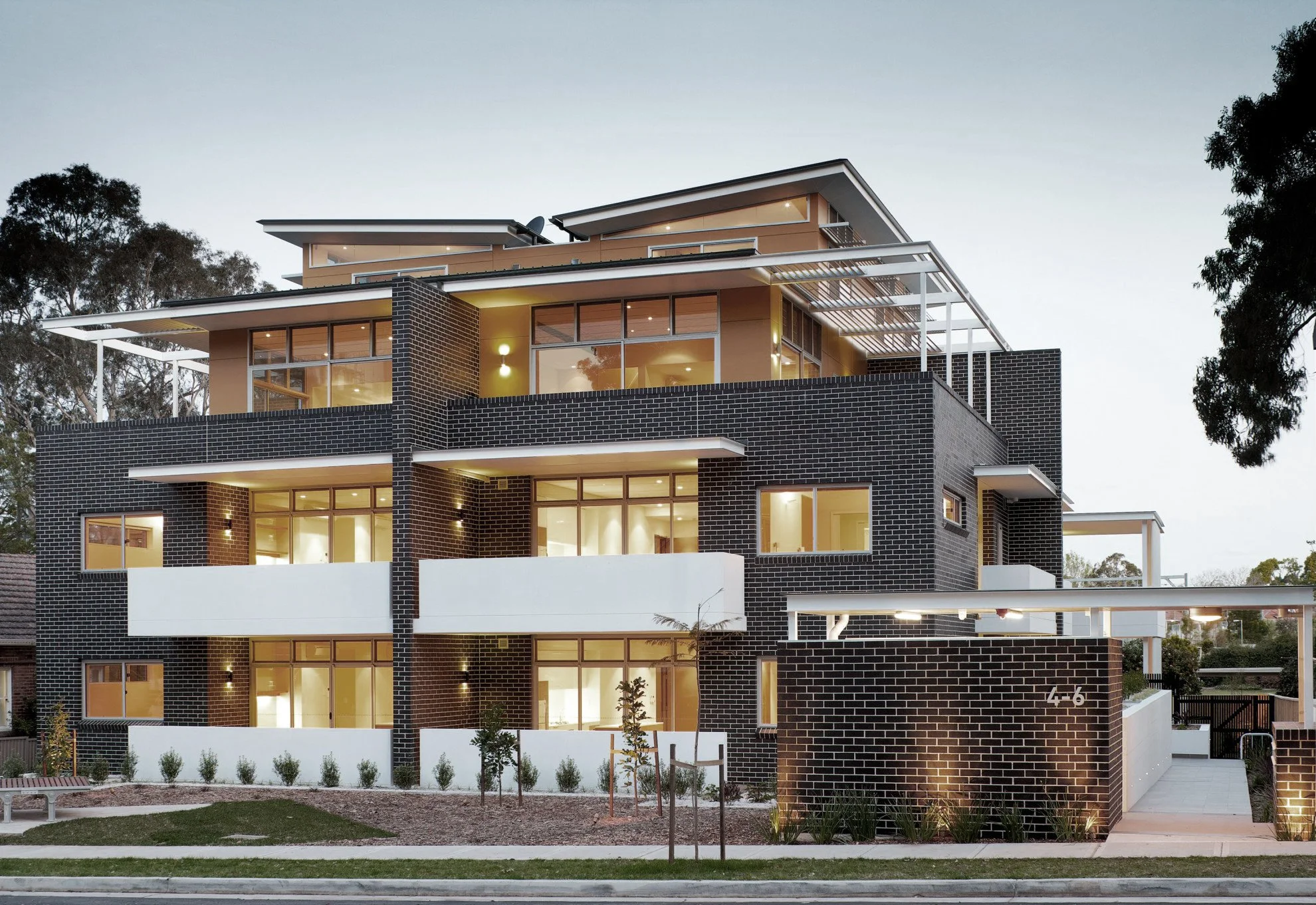 Modern multi-story residential building with black brick exterior, large glass windows, and outdoor balconies, illuminated in evening light.