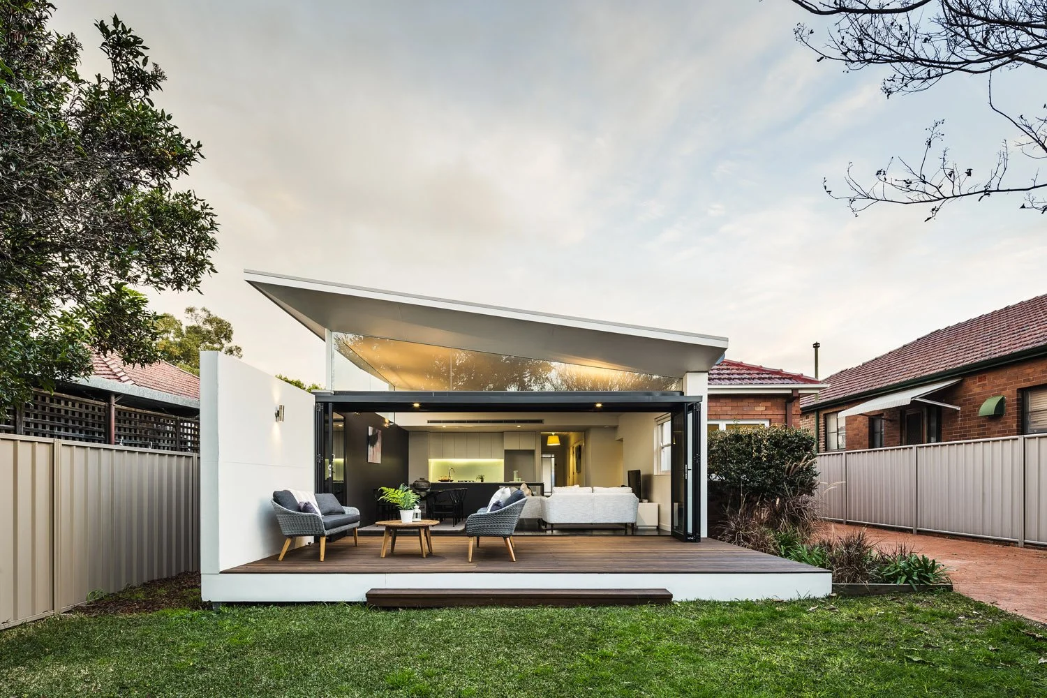 Modern backyard with an open living space, wooden deck, outdoor chairs, small table, and view into a cozy indoor area with sofas and tabletop plants, surrounded by a beige fence and neighboring houses, during early evening.