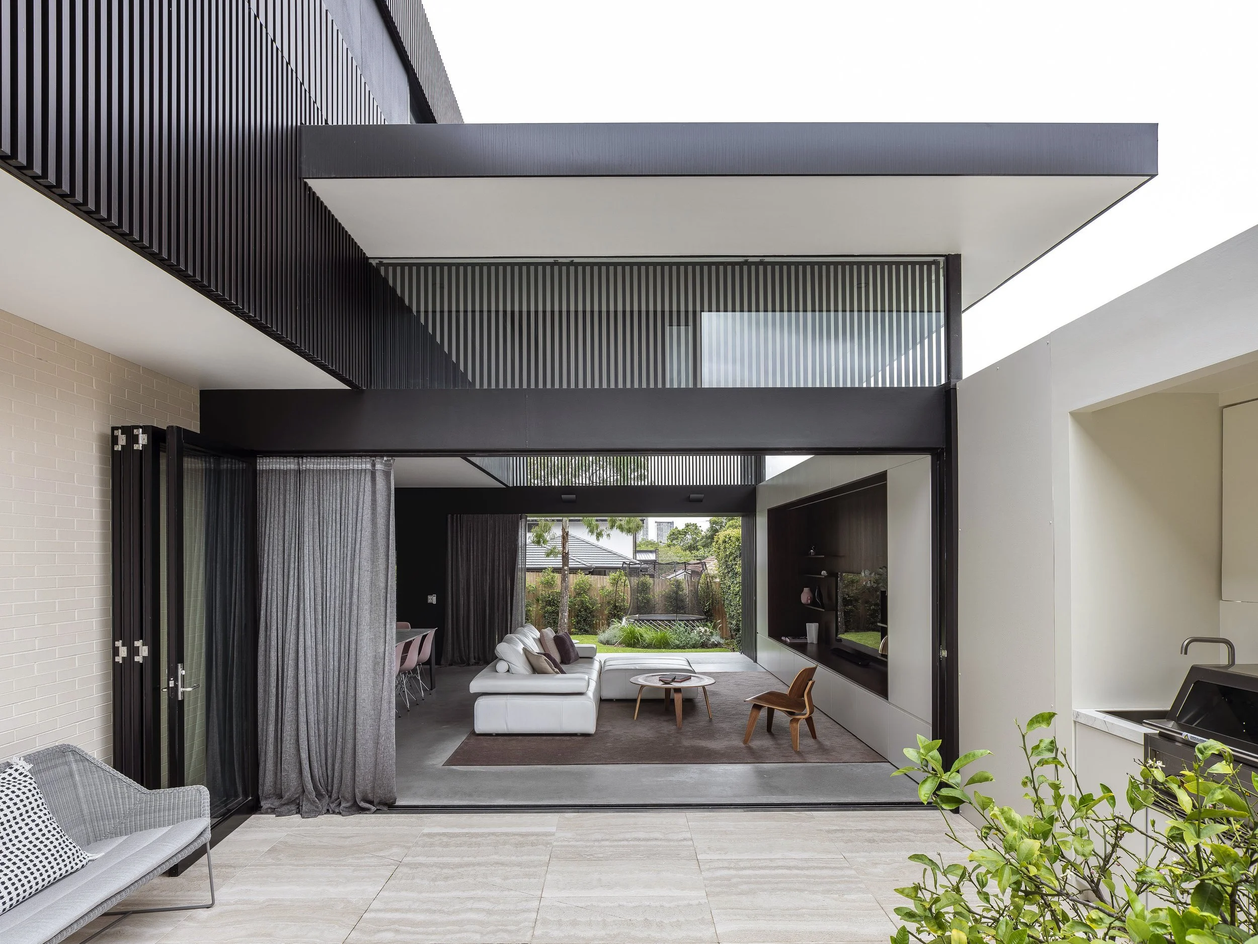 Modern living room visible through large sliding glass doors, with white sectional sofa, wooden chairs, and outdoor garden view.