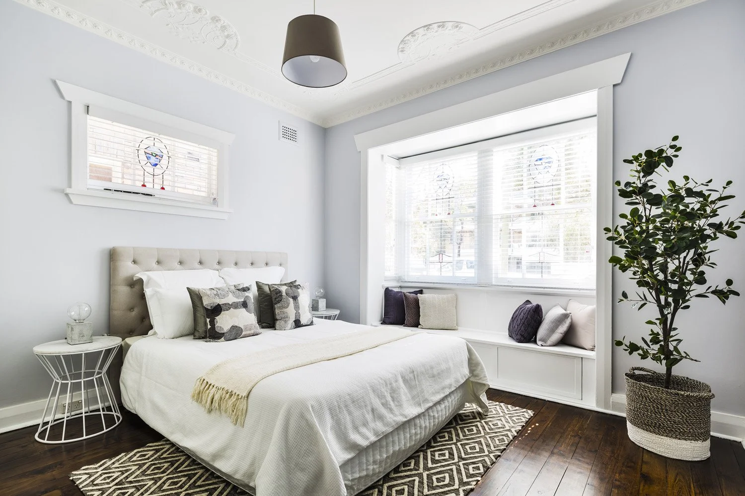 Bright bedroom with white walls and dark hardwood floors, a bed with white bedding and patterned pillows, a window seat with cushions, a potted plant, and a modern pendant light.
