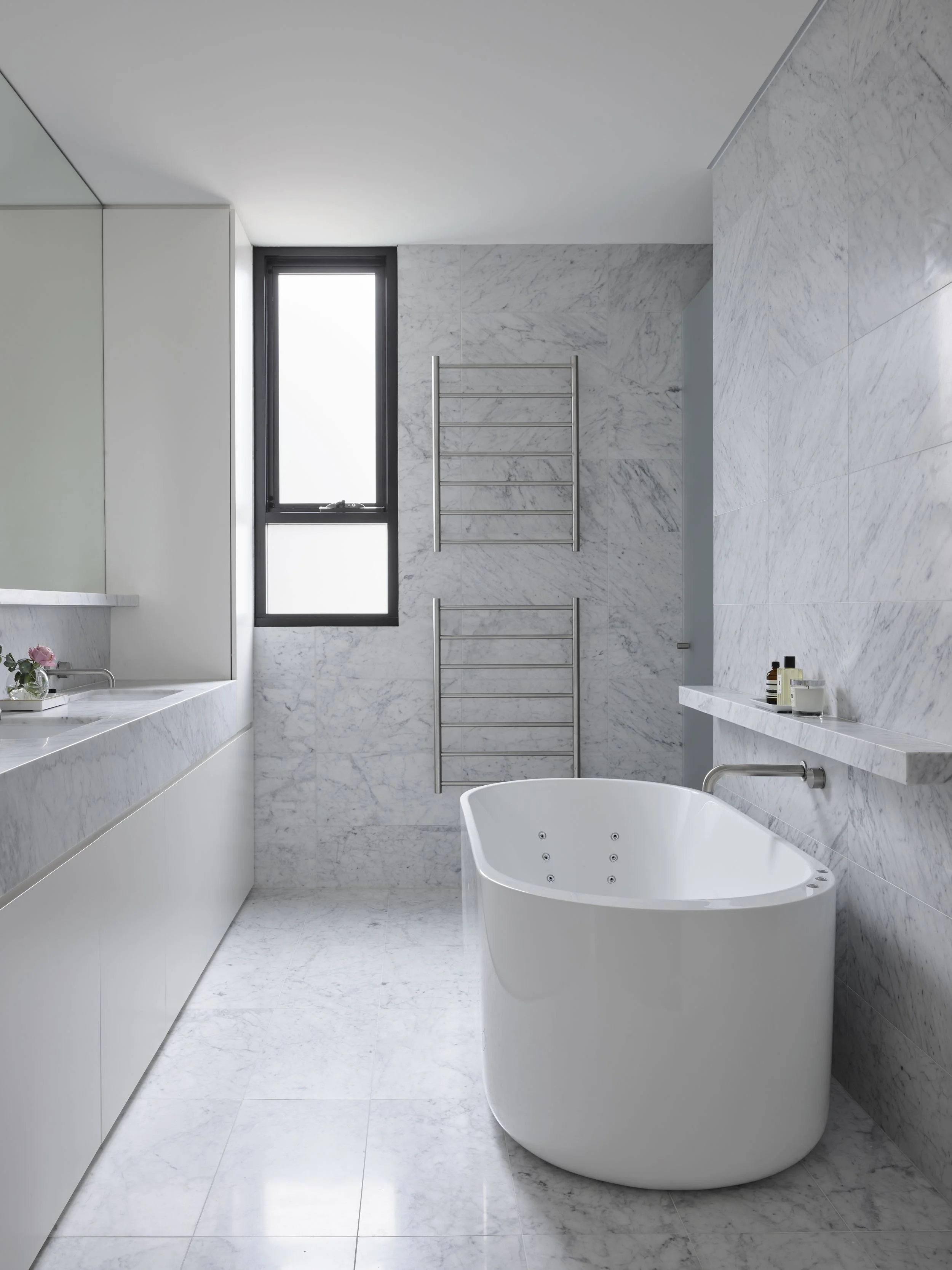 Modern bathroom with white marble walls and floor, a freestanding bathtub, a large mirror, a window, and towel racks.