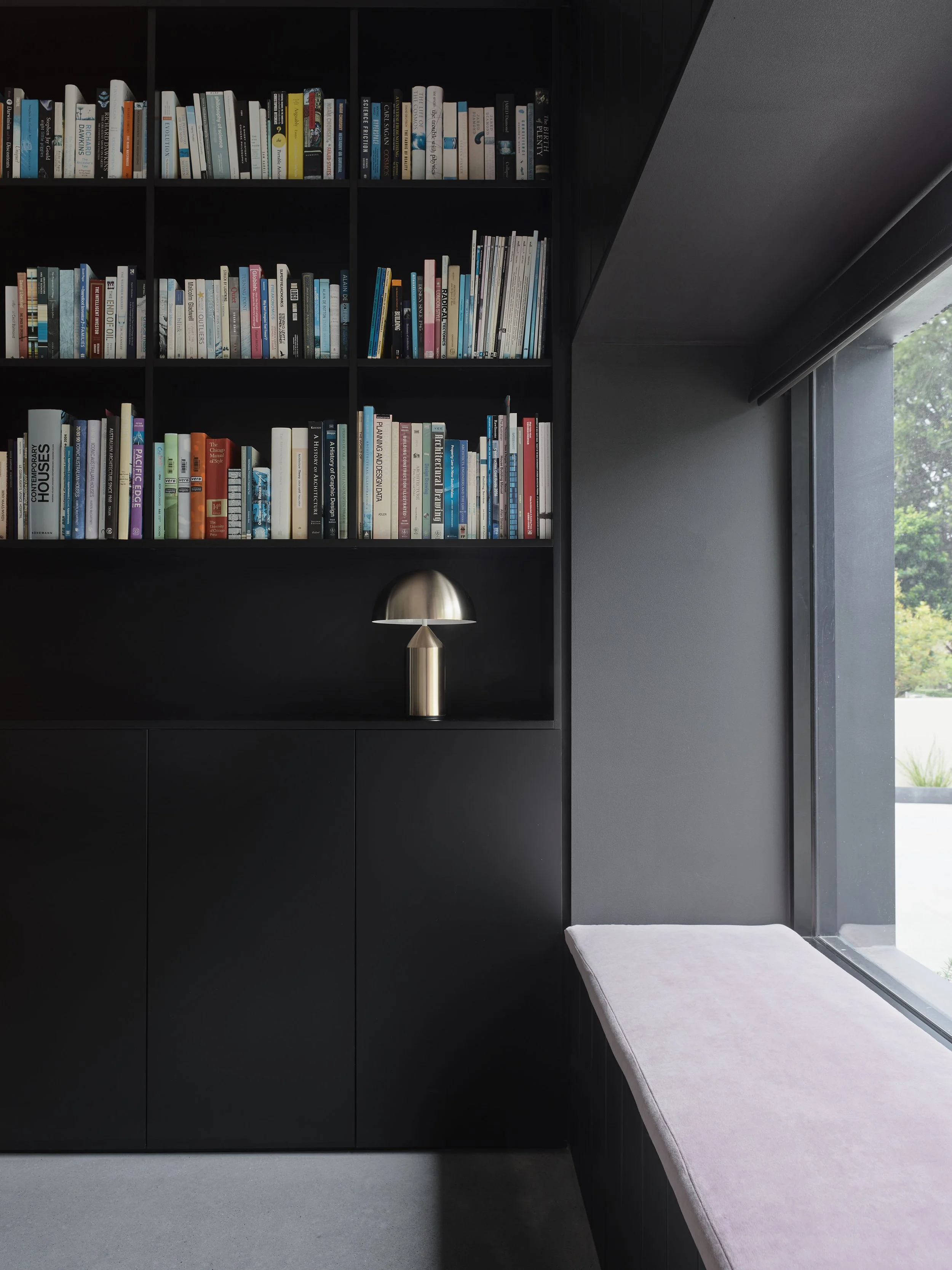 A black bookshelf filled with books next to a window with a pink cushioned bench underneath.