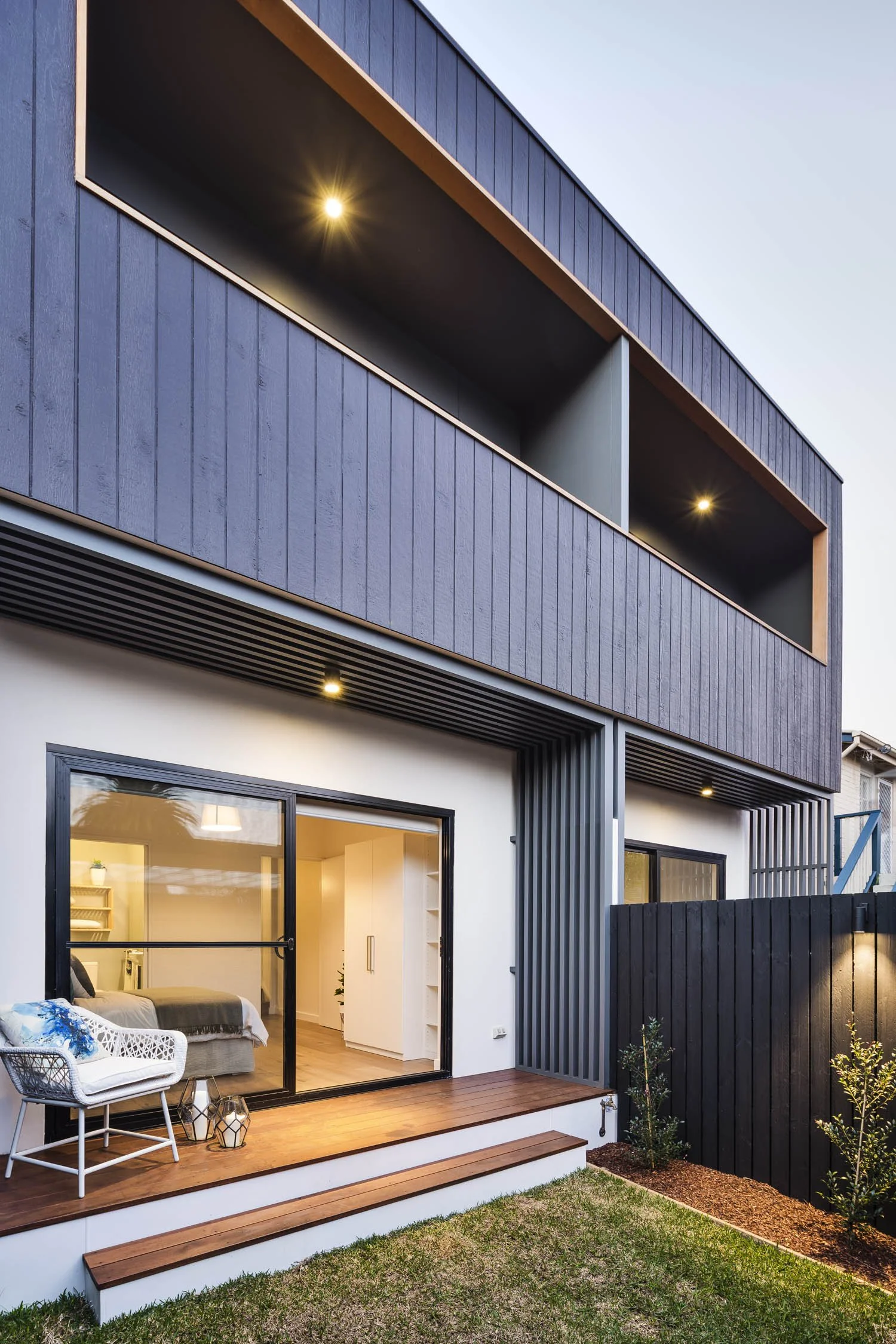 Modern two-story house with a backyard and a patio, featuring sliding glass doors, a white interior with a bed, and outdoor seating with lanterns.